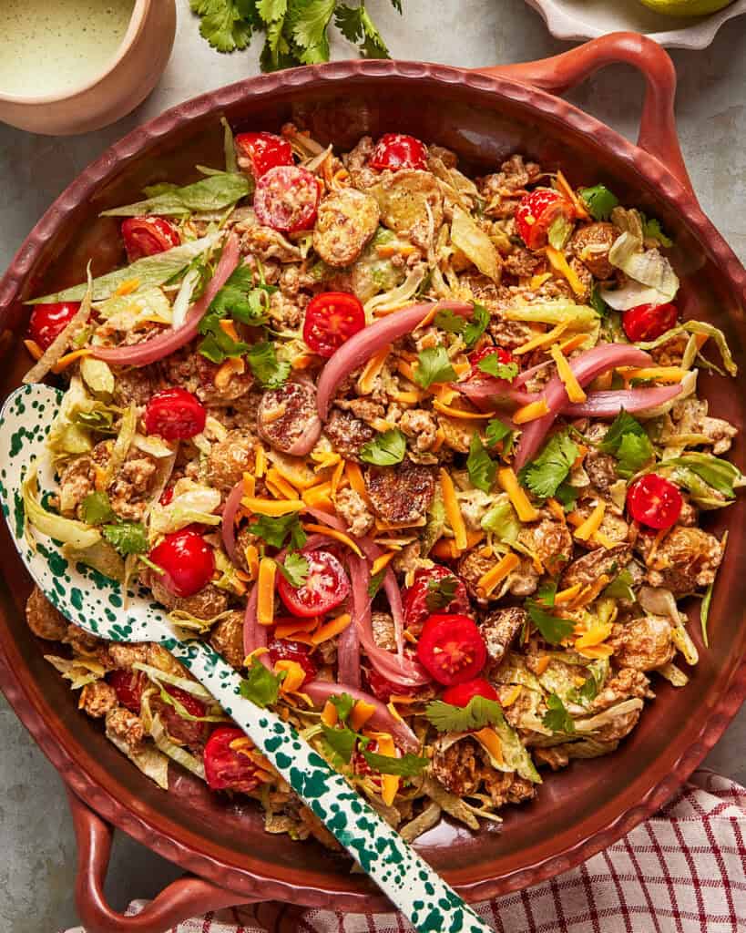 A large salad bowl filled with chopped lettuce, cherry tomatoes, shredded cheddar cheese, pickled onions, crispy tortilla strips, fresh cilantro, and grilled chicken, with a serving spoon resting inside.