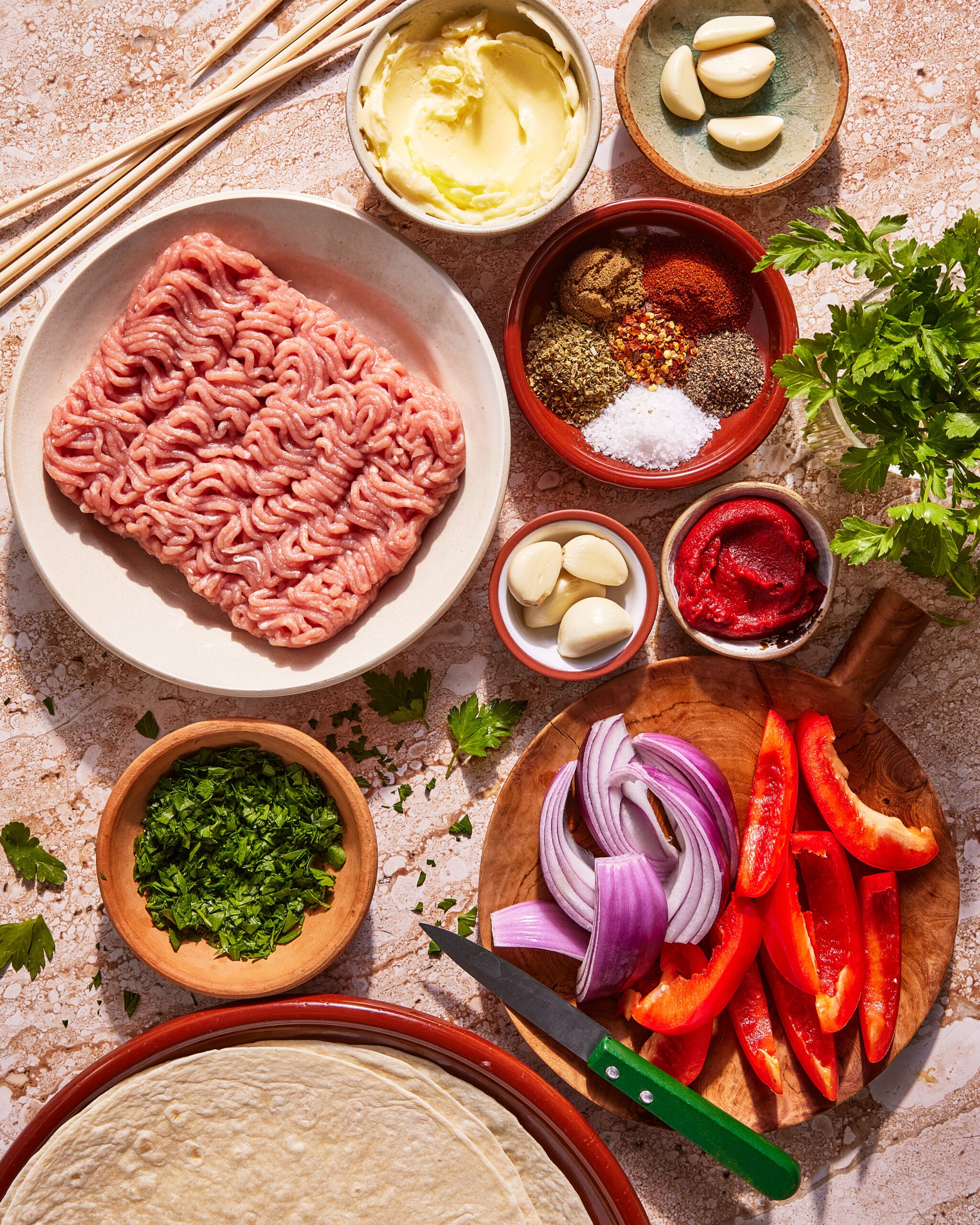 Top-down view of ingredients for a Middle Eastern dish: ground meat, tortilla wraps, sliced red onion, red bell pepper, fresh herbs, tomato paste, garlic, various spices, butter, and parsley on a stone surface.