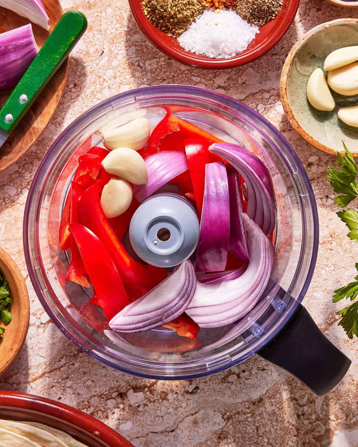 A food processor bowl containing red bell pepper slices, red onion wedges, and garlic cloves, surrounded by bowls of seasonings, herbs, and garlic on a beige countertop.