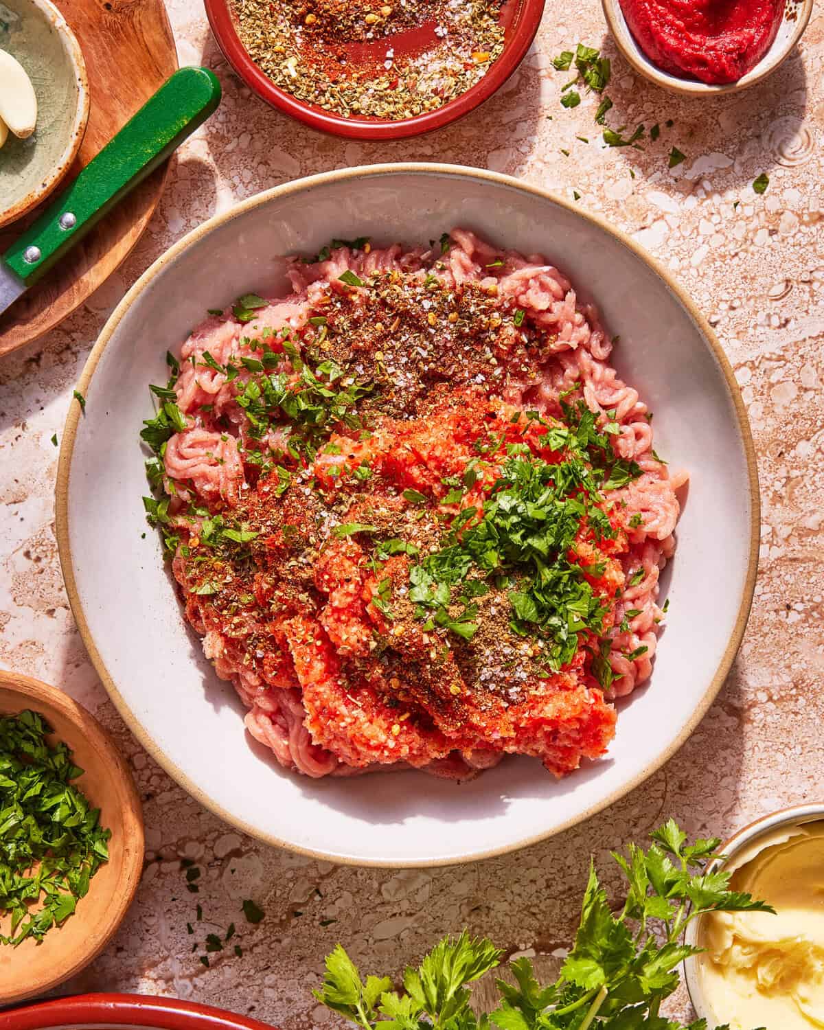 A bowl of raw ground meat topped with chopped parsley, crushed tomatoes, and various spices sits on a table surrounded by small bowls of herbs, tomato paste, and seasonings.