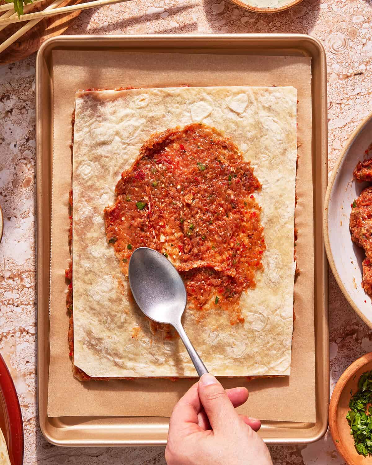A hand spreads tomato sauce onto a rectangular sheet of flatbread or lasagna pasta with a spoon, on a parchment-lined baking tray. Bowls of sauce and herbs are visible nearby.