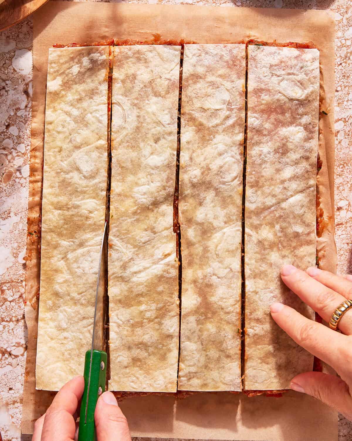 A person uses a green-handled knife to cut a large rectangular flatbread or pastry into four long strips on a sheet of parchment paper.