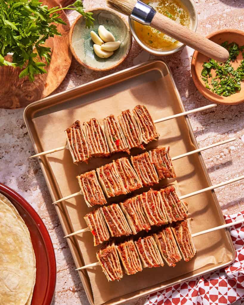 Four skewers with stacked, thin layers of seasoned meat or tortillas are arranged on a baking sheet lined with parchment paper. Surrounding the tray are bowls of fresh herbs, garlic, sauce, and uncooked tortillas.