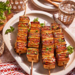 Four wooden skewers with grilled tofu and vegetables are arranged on a white platter, garnished with parsley. Two glasses, a small bowl of sauce, and forks are nearby on a beige and red-patterned tablecloth.