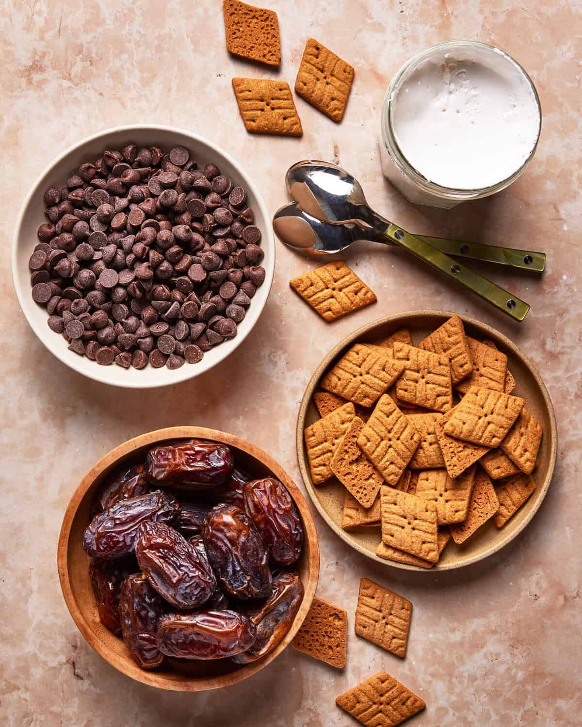 A top-down view of chocolate chips in a bowl, a bowl of whole dates, a plate of square graham crackers, a glass of milk, and two spoons on a light marble surface.