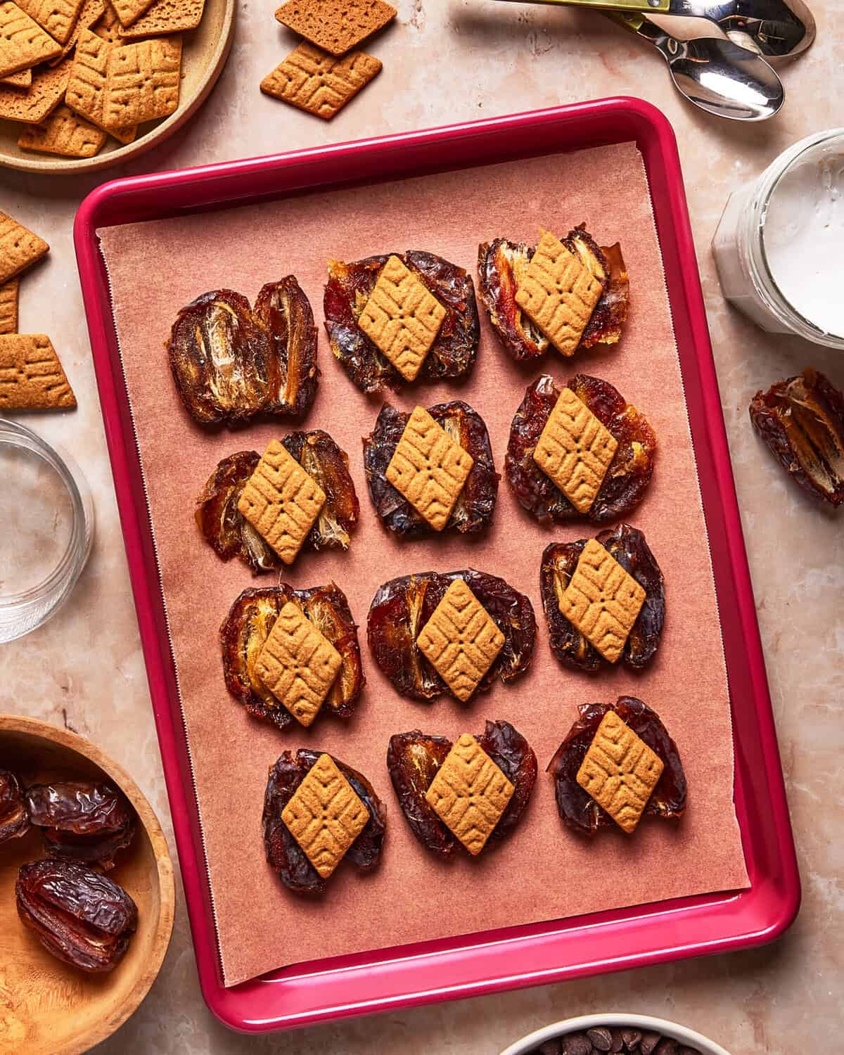 A red baking sheet lined with parchment displays dates stuffed with nut butter and topped with square graham cracker pieces. Bowls of dates, graham crackers, and a jar of white spread are nearby on a marble surface.