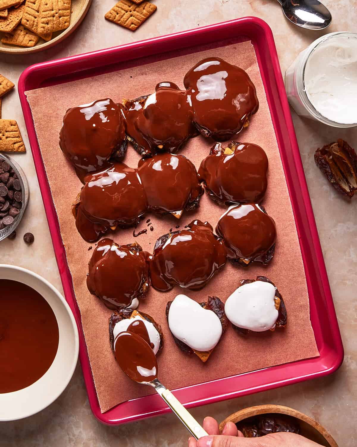 A tray lined with parchment paper holds snacks topped with melted chocolate and marshmallow. A hand uses a spoon to add marshmallow to a snack. Surrounding the tray are graham crackers, chocolate chips, and a bowl of melted chocolate.