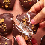 A person pulls apart a chocolate-covered treat revealing gooey marshmallow inside. The treat is topped with crumbled cookies and rests on a brown surface with more similar treats nearby.