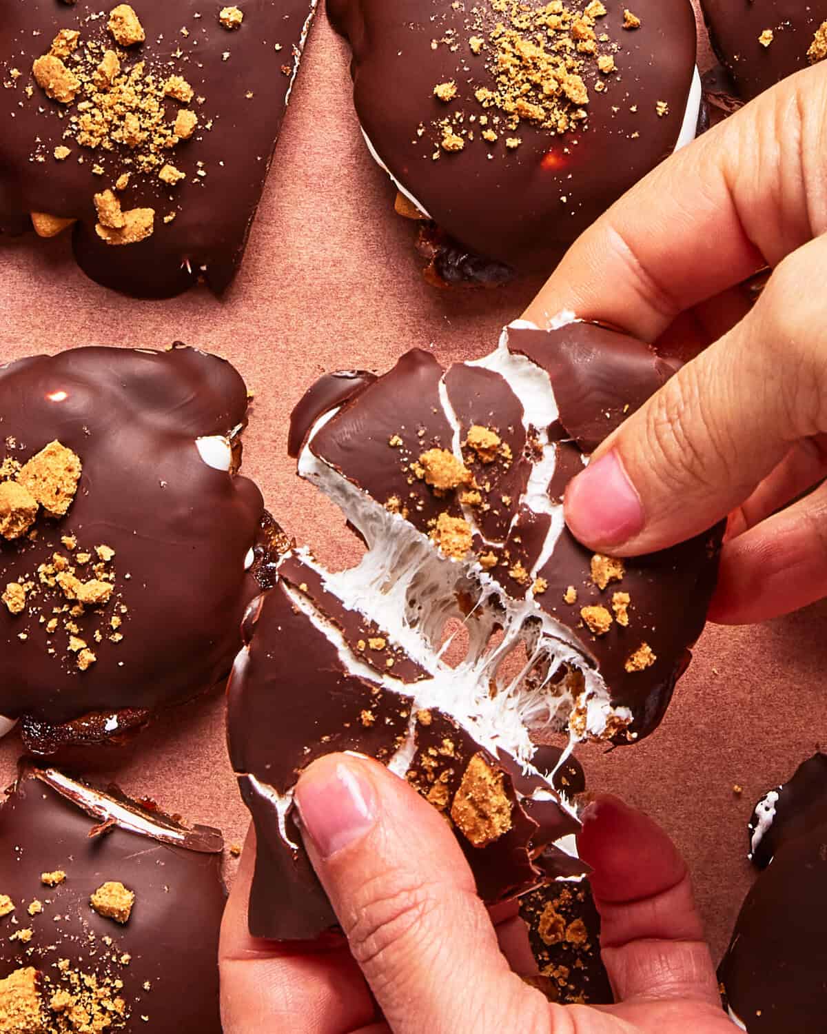 A person pulls apart a chocolate-covered treat revealing gooey marshmallow inside. The treat is topped with crumbled cookies and rests on a brown surface with more similar treats nearby.