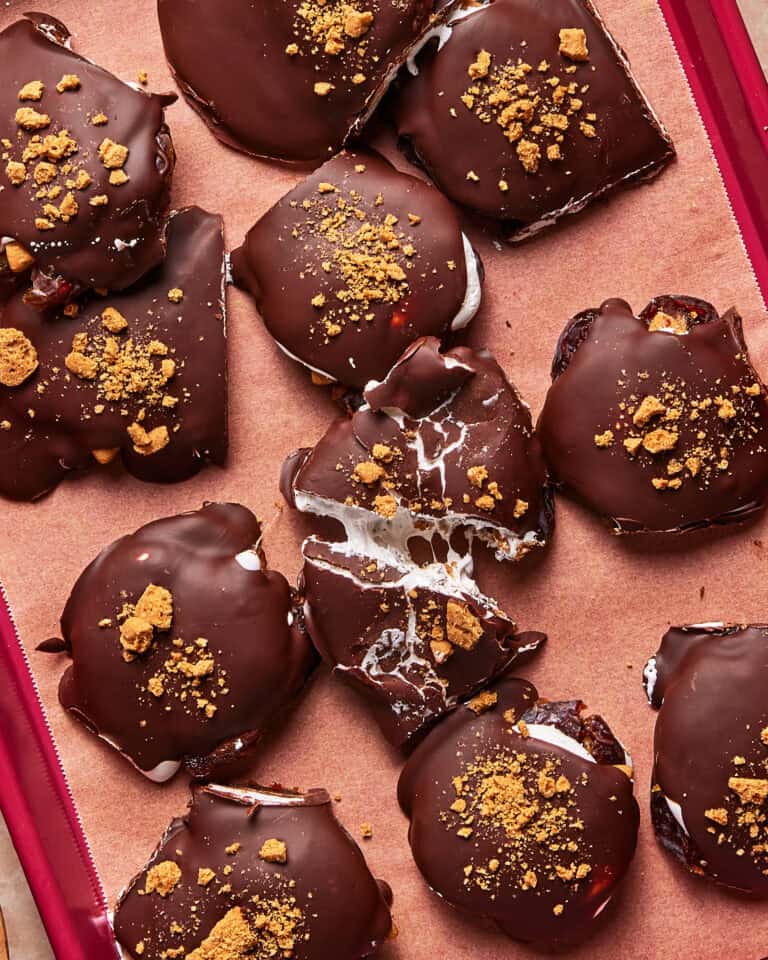 Chocolate-coated marshmallow and graham cracker treats are arranged on parchment paper, with some cracked open to reveal gooey filling. Crushed graham cracker pieces are sprinkled on top.