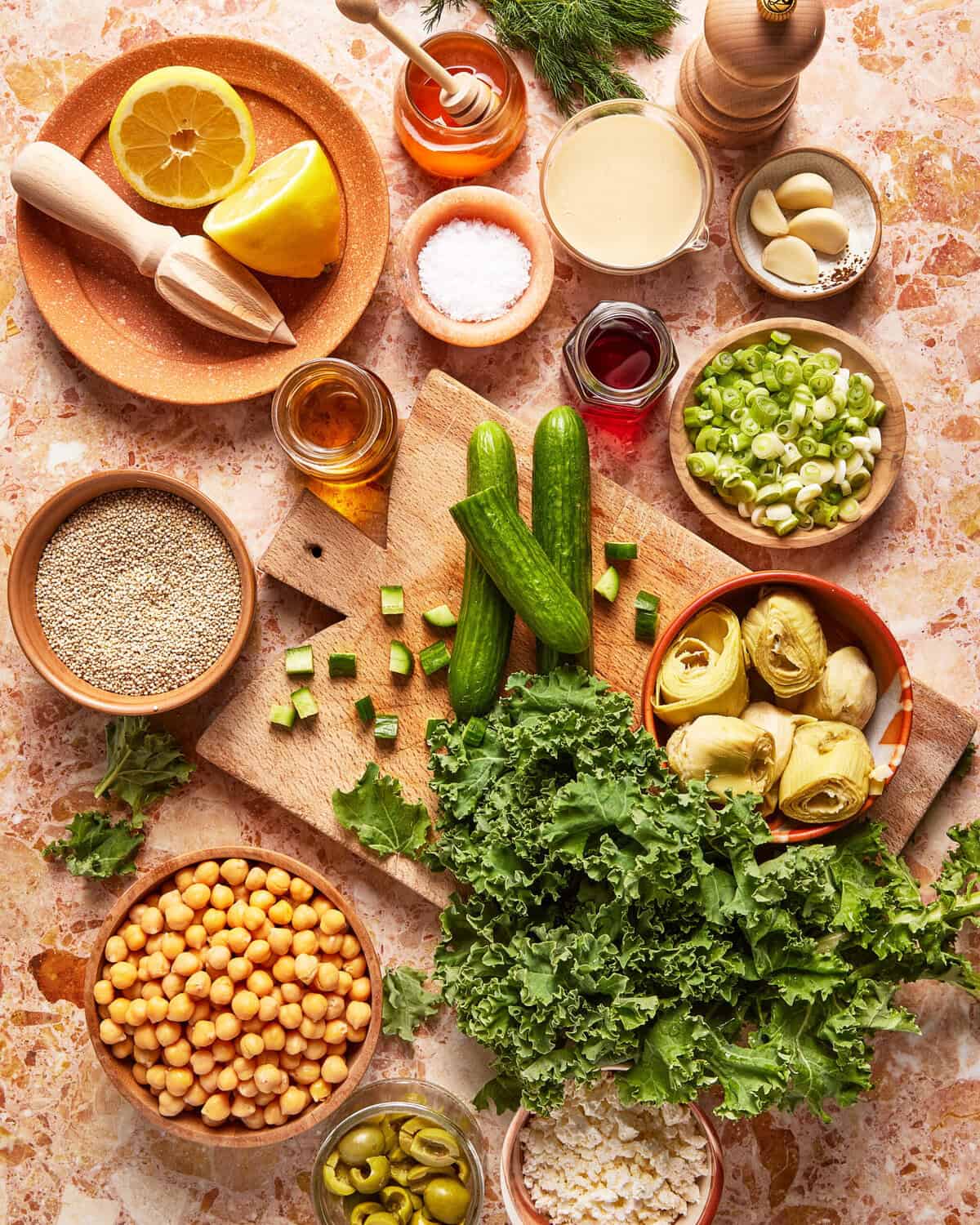 Various fresh ingredients on a countertop, including kale, cucumbers, chickpeas, artichoke hearts, green onions, olives, quinoa, garlic cloves, lemon, tahini, oats, and seasonings in small bowls.