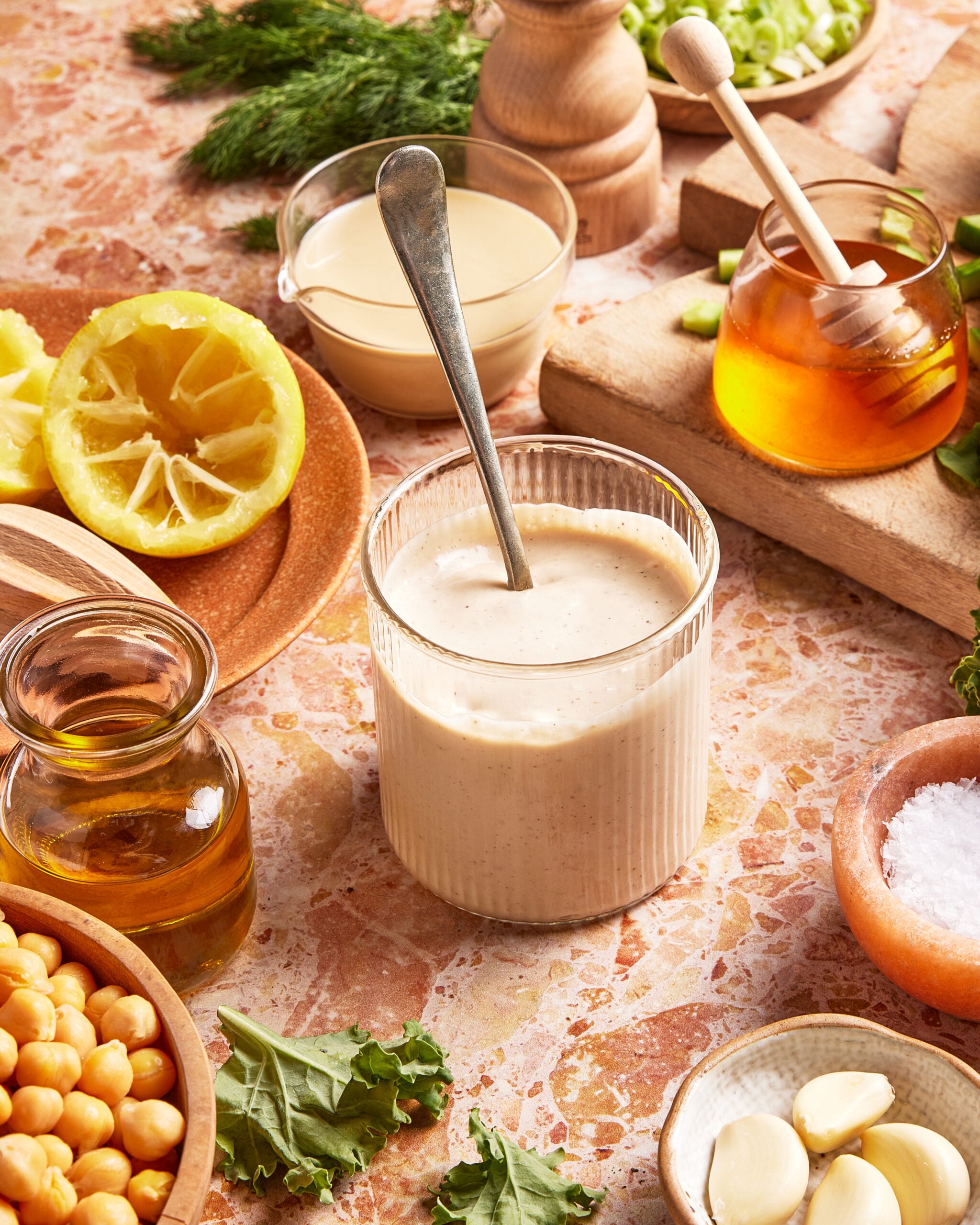 A glass of creamy dressing with a spoon inside, surrounded by lemon, honey, chickpeas, olive oil, salt, garlic, greens, fresh dill, and a bowl of tahini on a marble countertop.