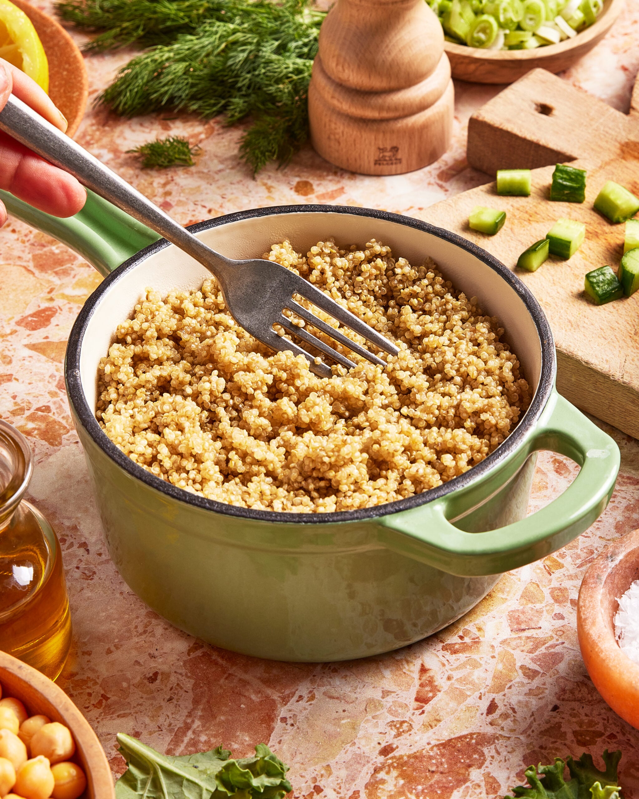A hand fluffs cooked quinoa with a fork in a green pot on a countertop, surrounded by chopped vegetables, fresh herbs, olive oil, and a pepper grinder.
