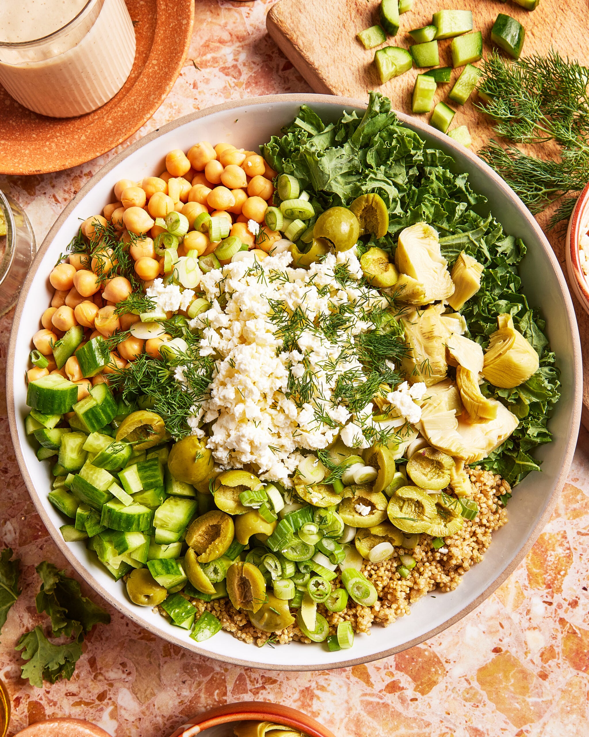 A large bowl filled with chickpeas, kale, cucumber, green olives, artichoke hearts, green onions, quinoa, feta cheese, and fresh dill arranged in colorful sections.
