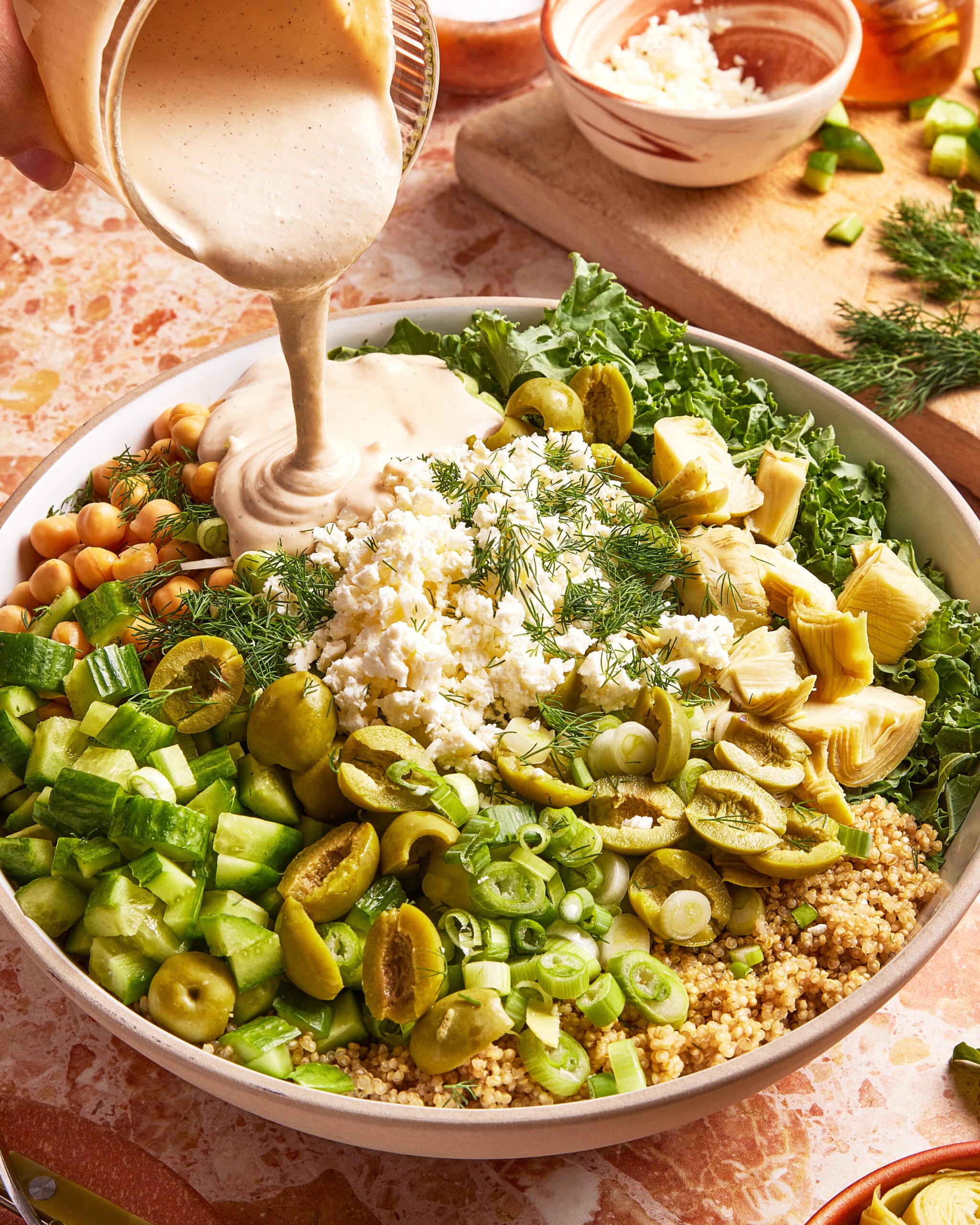 A large bowl filled with couscous, chopped cucumbers, green olives, artichokes, chickpeas, kale, feta cheese, and fresh herbs, with creamy dressing being poured on top.