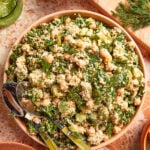 A bowl of chickpea salad with quinoa, chopped greens, herbs, feta cheese, and cucumber, served with salad tongs on a marble surface. Other ingredients and a green dressing are visible nearby.