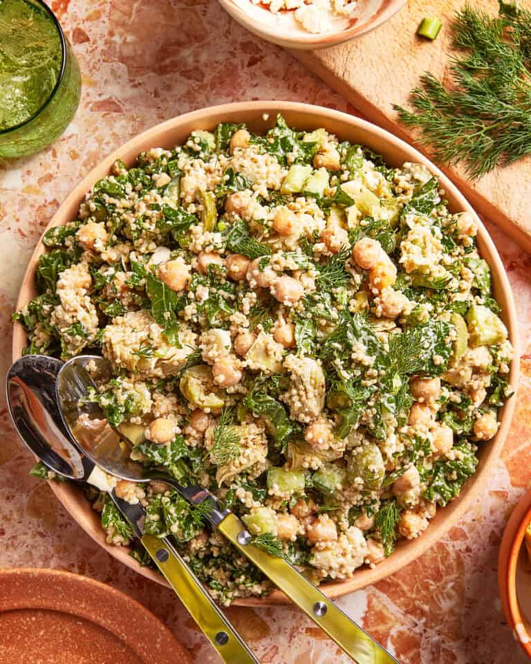 A bowl of chickpea salad with quinoa, chopped greens, herbs, feta cheese, and cucumber, served with salad tongs on a marble surface. Other ingredients and a green dressing are visible nearby.