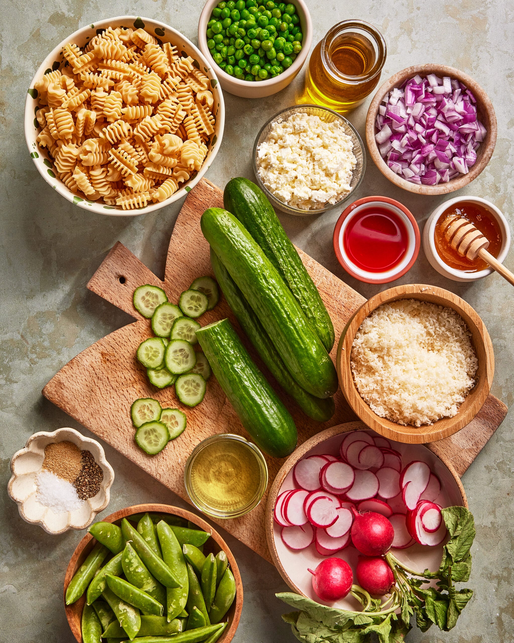 An overhead view of ingredients on a table, including rotini pasta, cucumbers, radishes, snap peas, peas, chopped red onion, grated cheese, crumbled cheese, honey, olive oil, red vinegar, salt, and pepper.