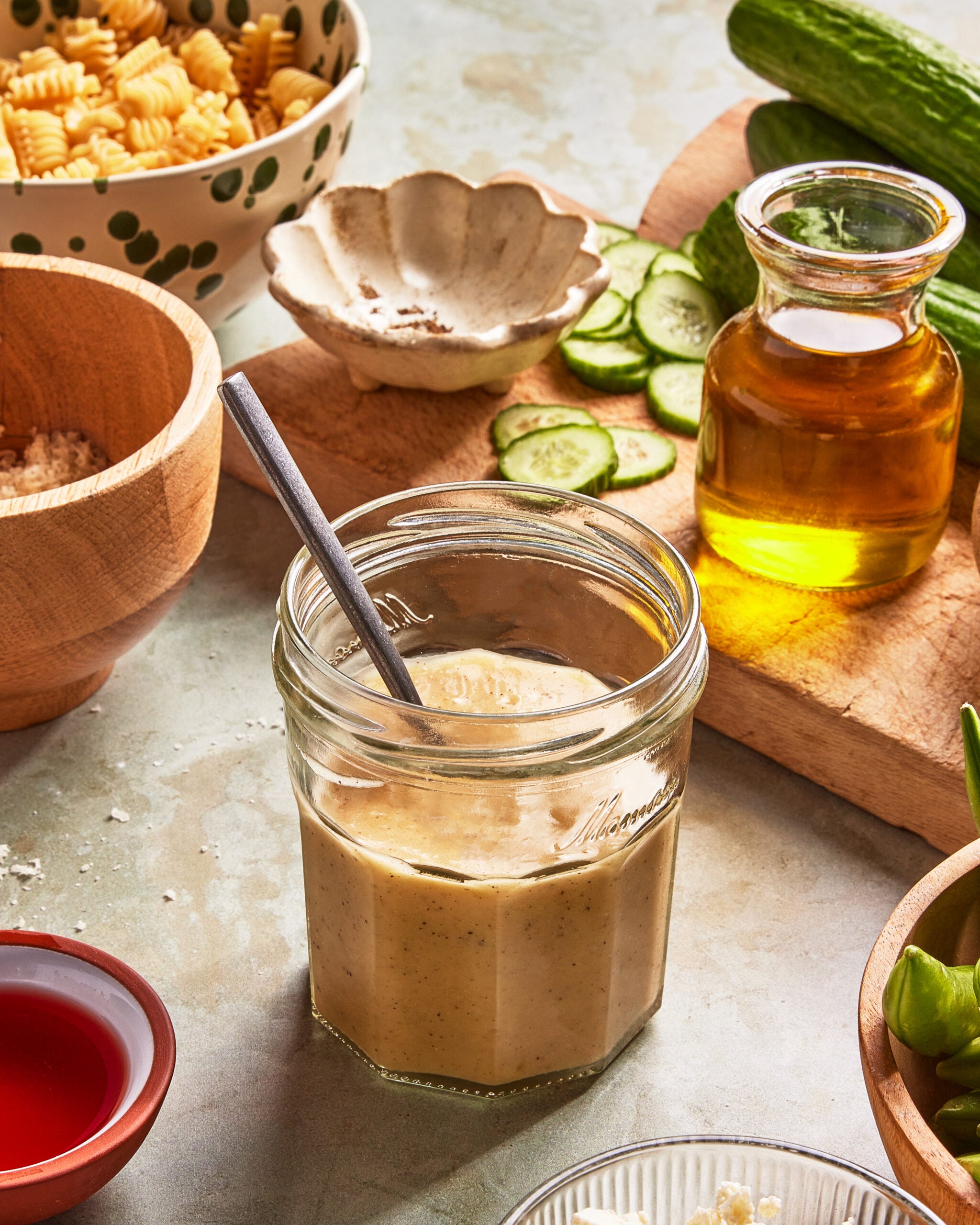 A jar of creamy dressing with a spoon inside, surrounded by a bowl of pasta, a small bottle of olive oil, sliced cucumbers on a wooden board, and various bowls containing ingredients on a light countertop.