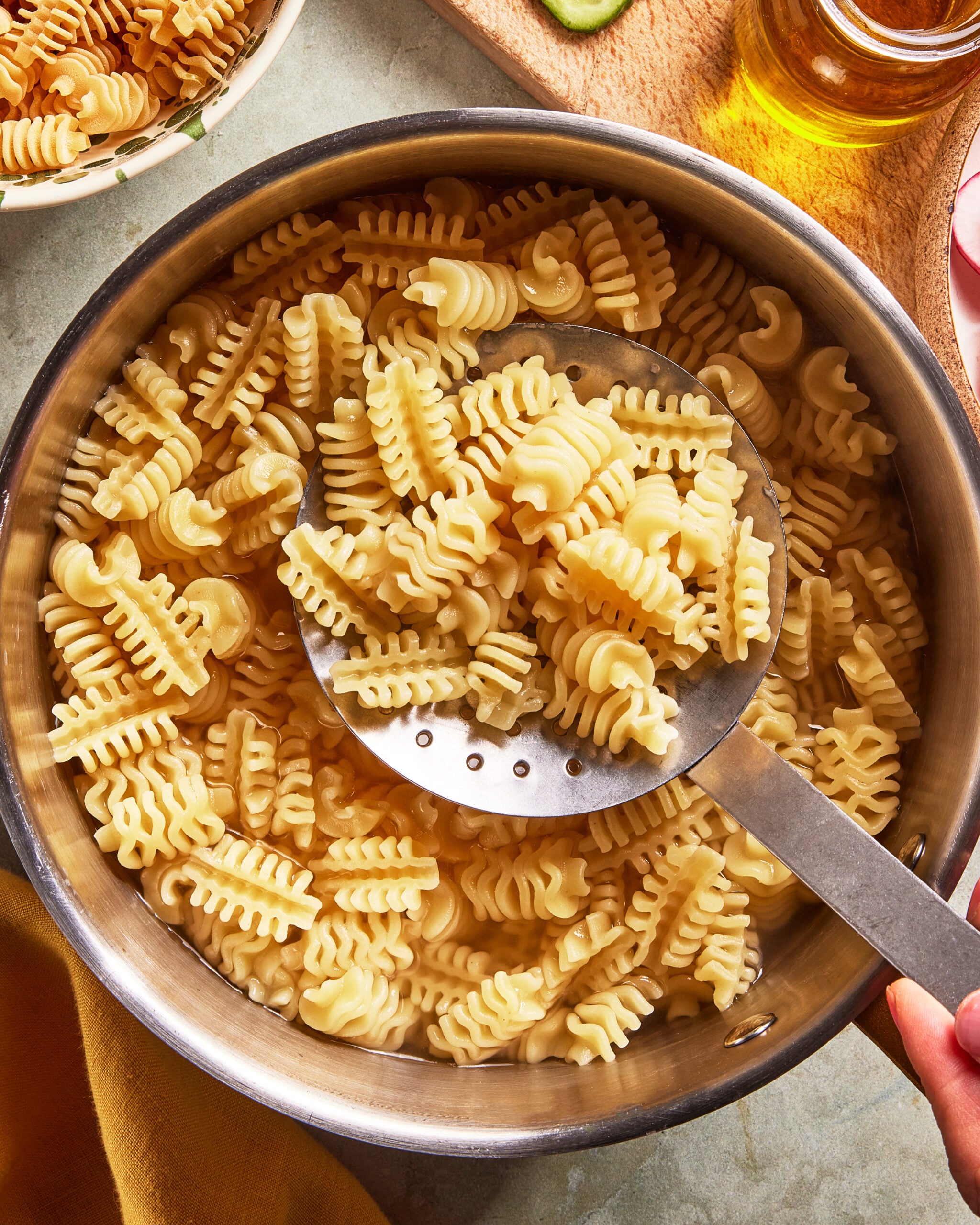 A metal pot filled with cooked mafaldine pasta, with a slotted spoon lifting some noodles. The pasta has a wavy, ruffled edge. The scene is brightly lit, with part of a dish, cutting board, and bottle visible nearby.
