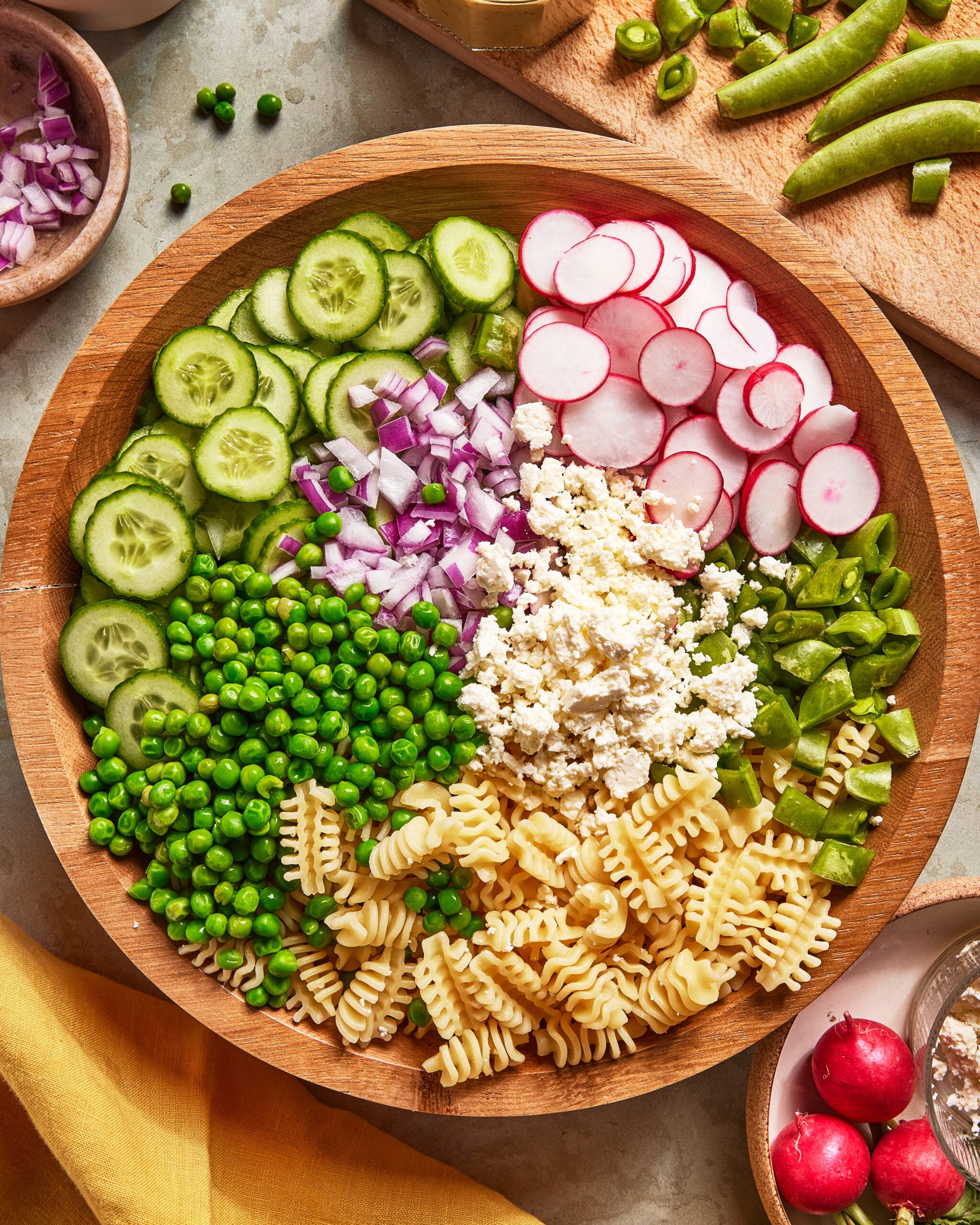 A wooden bowl filled with radiatori pasta, green peas, sliced cucumbers, chopped red onions, sliced radishes, snap peas, and crumbled cheese, arranged in sections. Cutting boards with veggies are nearby.