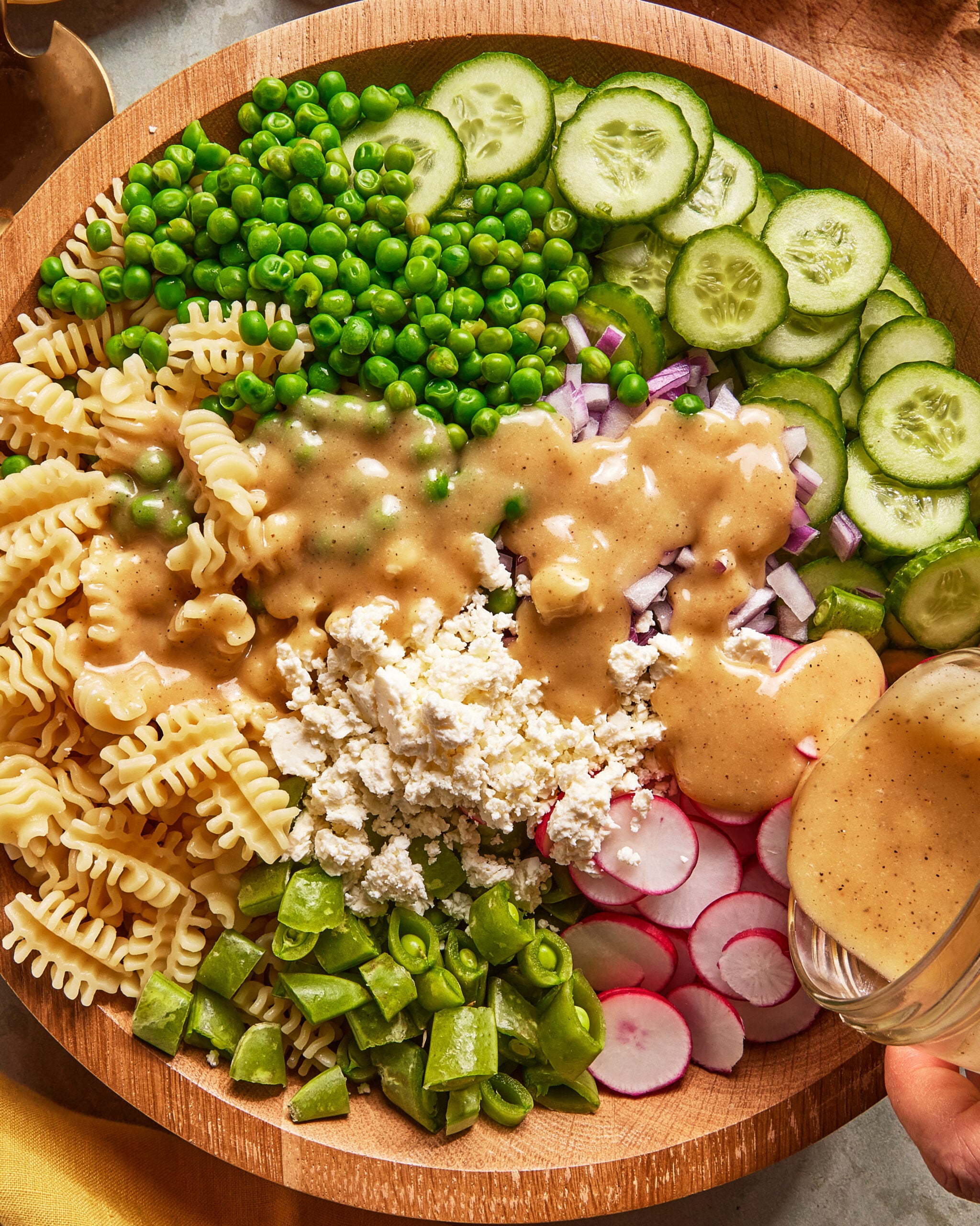 A wooden bowl filled with pasta, green peas, sliced cucumbers, chopped red onion, sliced radishes, snap peas, crumbled feta, and creamy dressing being poured over the salad.