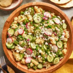 A wooden bowl filled with pasta salad containing rotini pasta, sliced cucumbers, radishes, red onion, green peas, asparagus, and crumbled feta cheese, set on a table with plates, cutlery, and a yellow napkin.