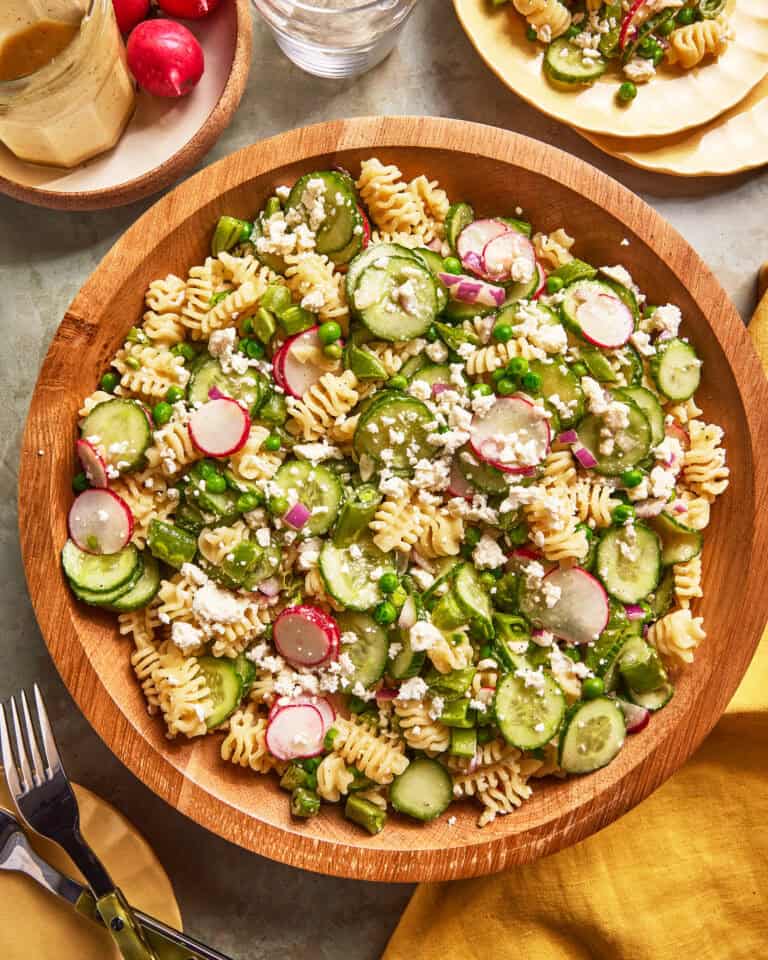 A wooden bowl filled with pasta salad containing rotini pasta, sliced cucumbers, radishes, red onion, green peas, asparagus, and crumbled feta cheese, set on a table with plates, cutlery, and a yellow napkin.