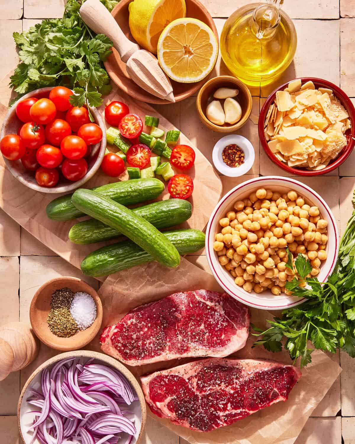 A top-down view of fresh ingredients on a counter, including raw steak, cherry tomatoes, cucumbers, chickpeas, red onion slices, lemon, garlic, parsley, cilantro, olive oil, shaved parmesan, and seasoning bowls.