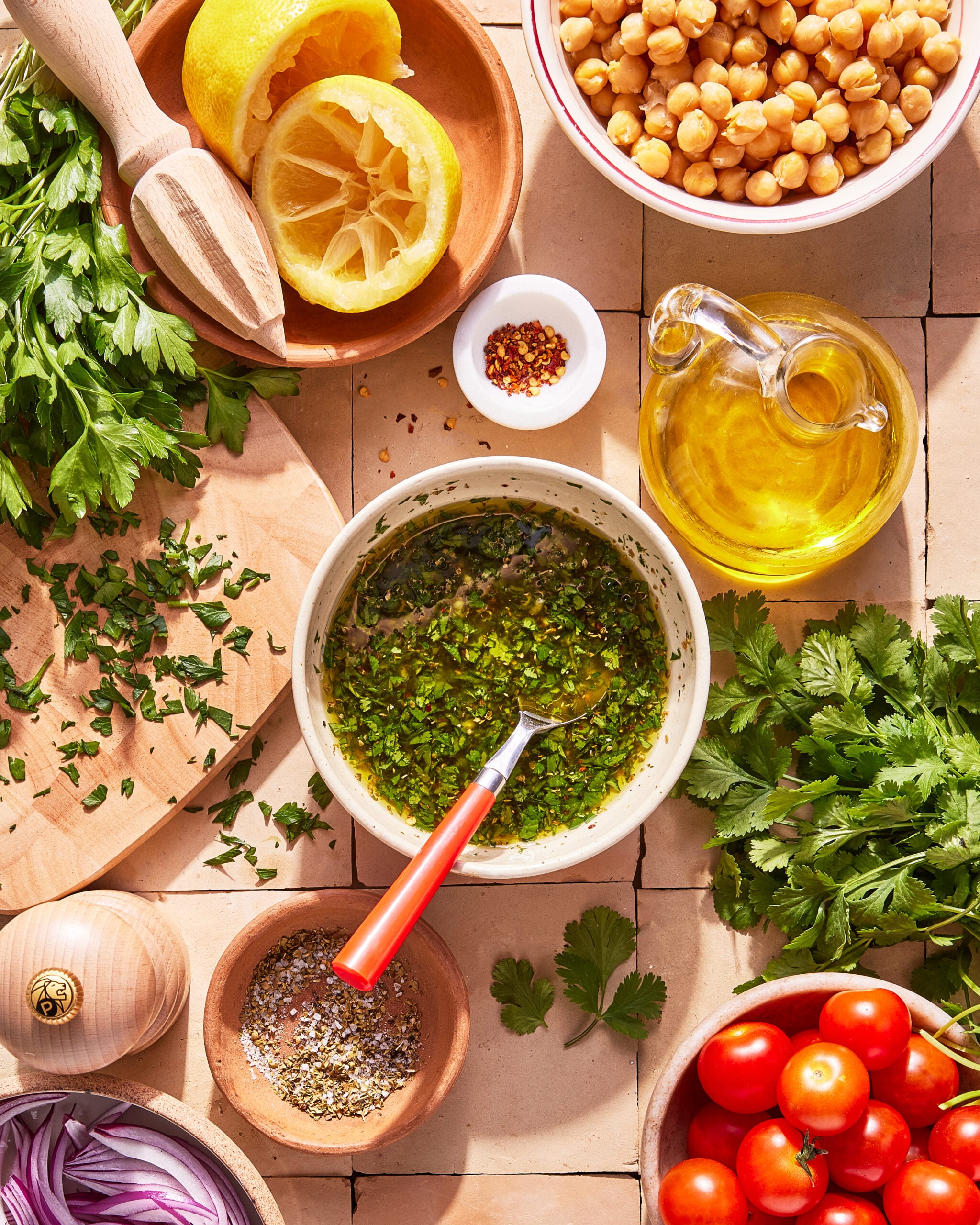 Overhead view of a table with chopped parsley, halved lemon, chickpeas, olive oil, cherry tomatoes, red onion, spices, and a bowl of herb dressing with a spoon, arranged on a tiled surface.