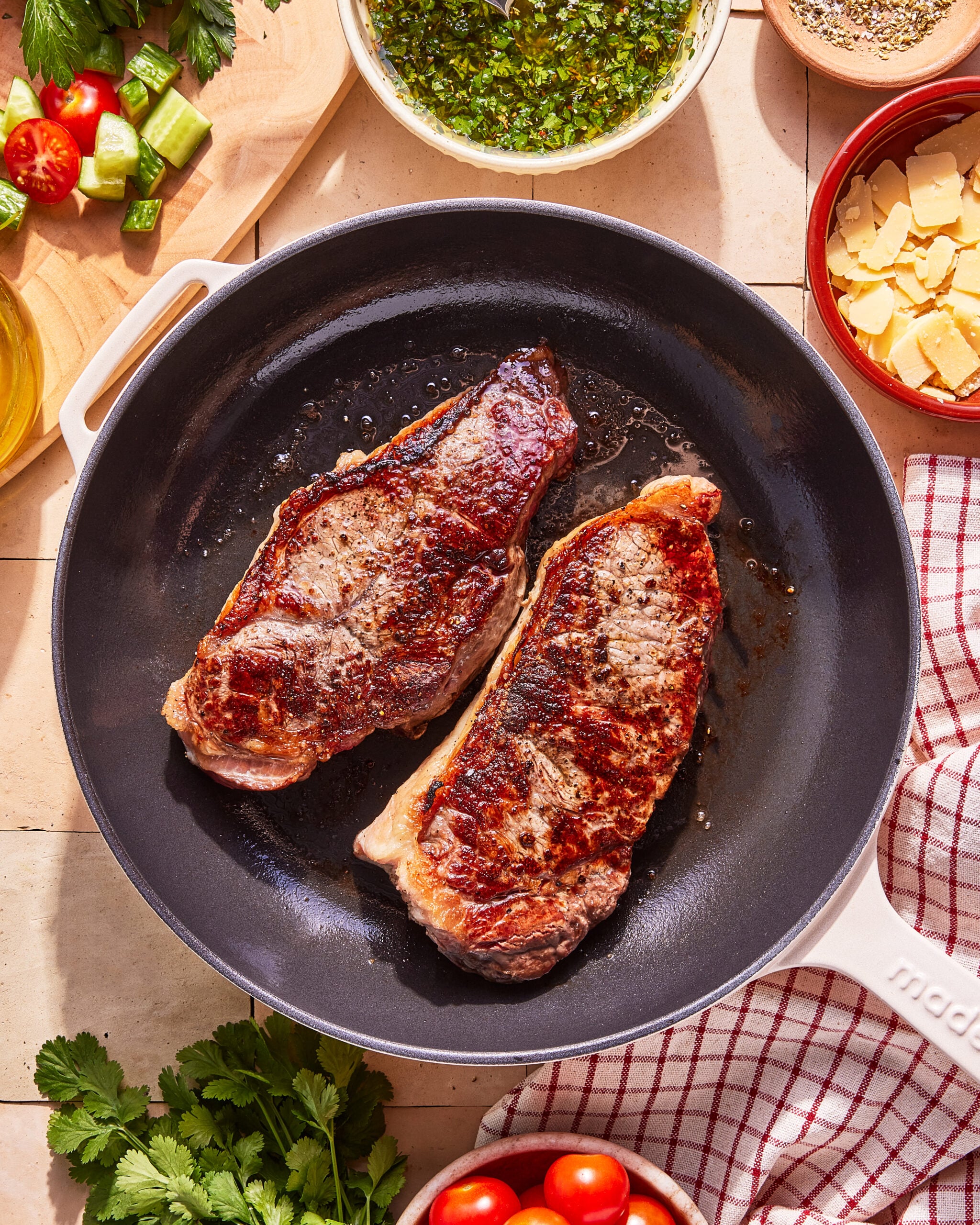 Two seared steaks cooking in a black skillet, surrounded by bowls of herbs, grated cheese, cherry tomatoes, cucumber, and fresh cilantro on a light-colored countertop.