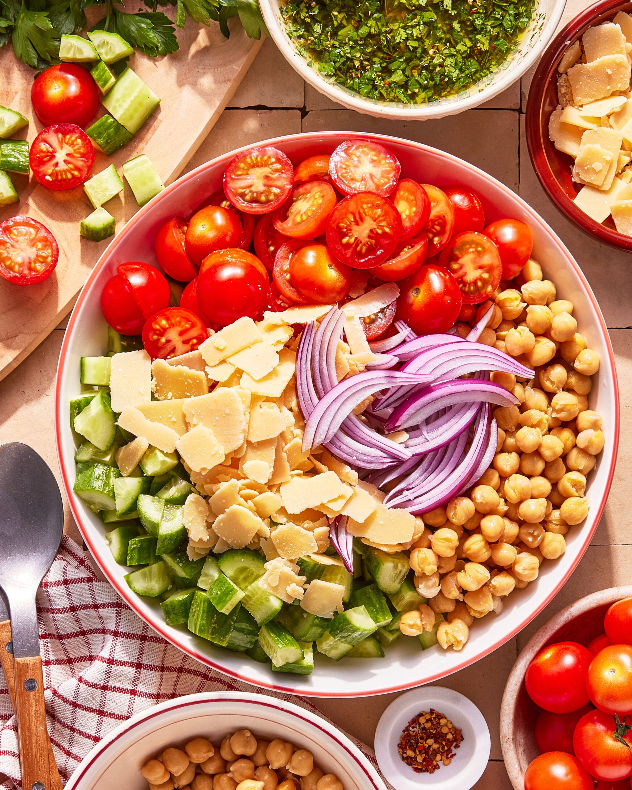 A bowl filled with chopped cherry tomatoes, cucumber, red onion, chickpeas, and shaved parmesan cheese, surrounded by more vegetables, a bowl of dressing, and kitchen utensils on a tiled surface.