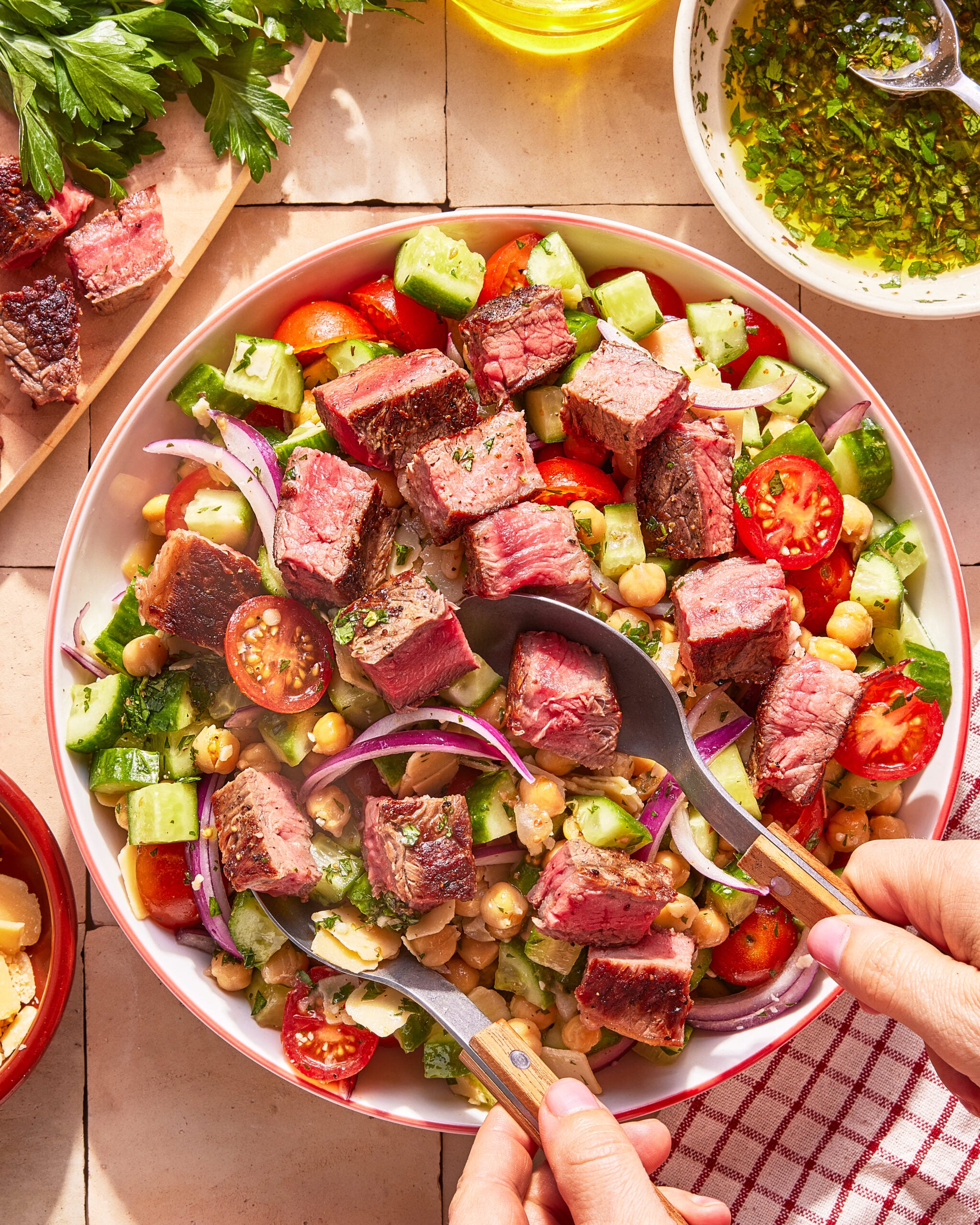 A bowl of salad with grilled steak cubes, cherry tomatoes, cucumber, red onion, chickpeas, and herbs being served with utensils; fresh parsley and a bowl of dressing are nearby.