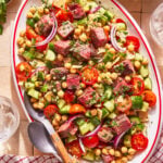 An oval platter of salad with grilled steak pieces, cherry tomatoes, cucumber, chickpeas, red onion, herbs, and cheese, served with a metal serving spoon and fork on a tiled surface next to a checkered cloth.
