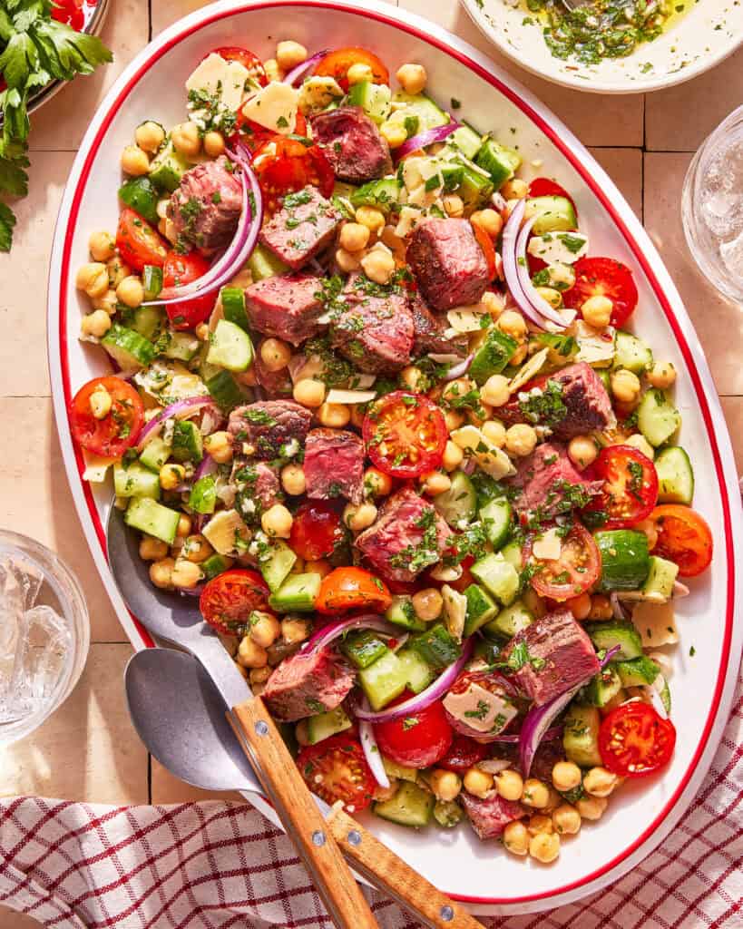 An oval platter of salad with grilled steak pieces, cherry tomatoes, cucumber, chickpeas, red onion, herbs, and cheese, served with a metal serving spoon and fork on a tiled surface next to a checkered cloth.