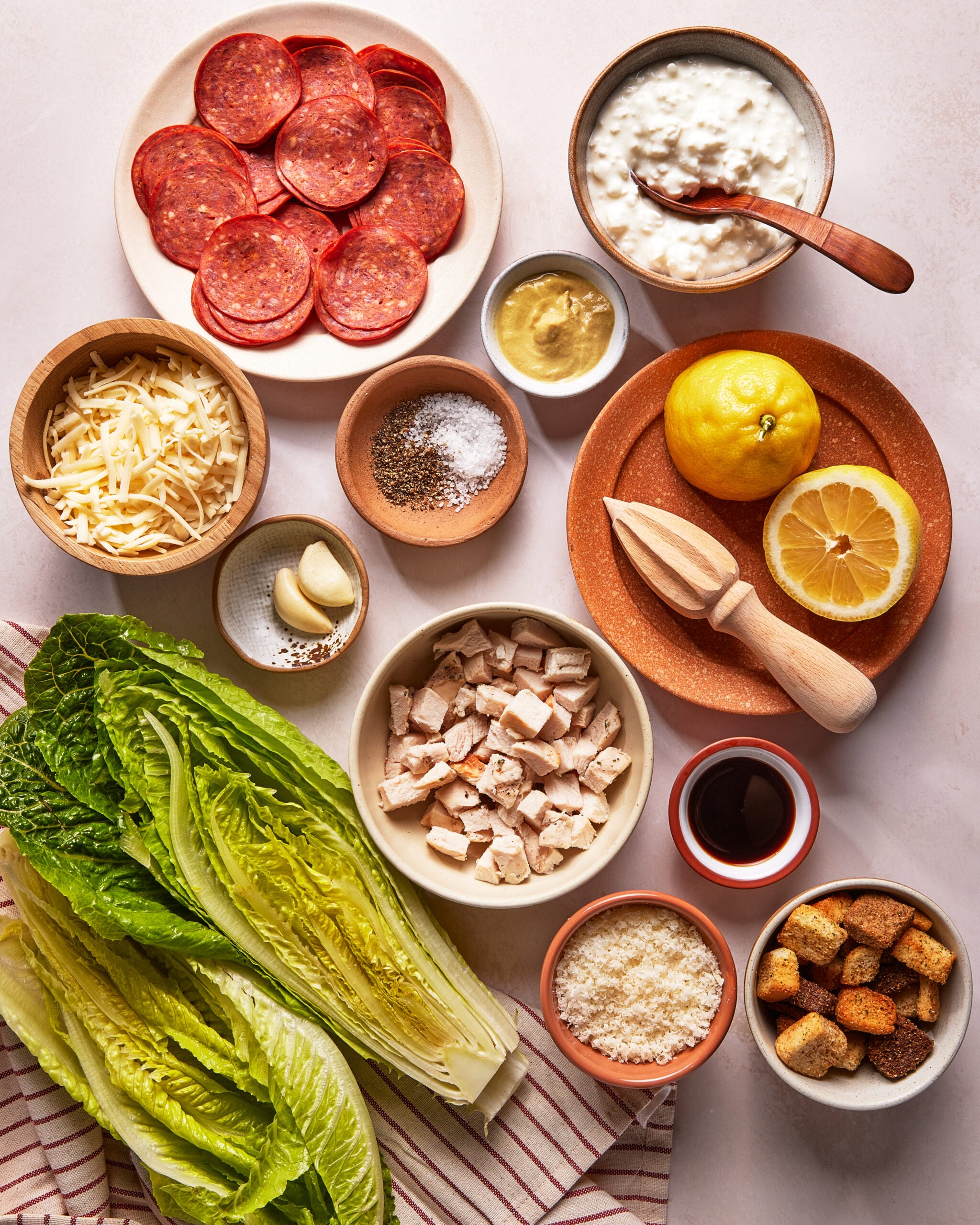 Ingredients for a salad arranged on a table, including romaine lettuce, sliced pepperoni, cubed chicken, shredded cheese, lemon, garlic, cottage cheese, mustard, croutons, Parmesan, spices, and Worcestershire sauce.