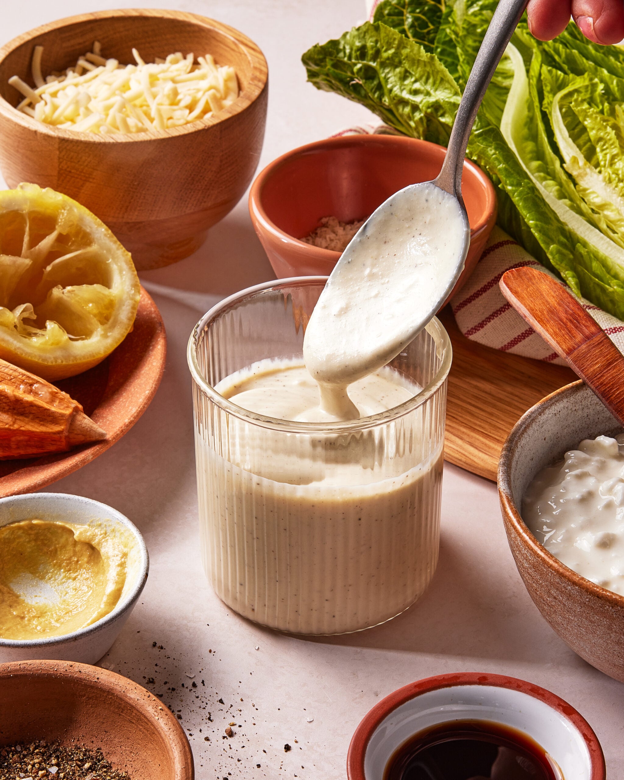 A spoon pours creamy Caesar salad dressing into a glass, surrounded by ingredients like grated cheese, lettuce, lemon, mustard, black pepper, and a bowl of cottage cheese.