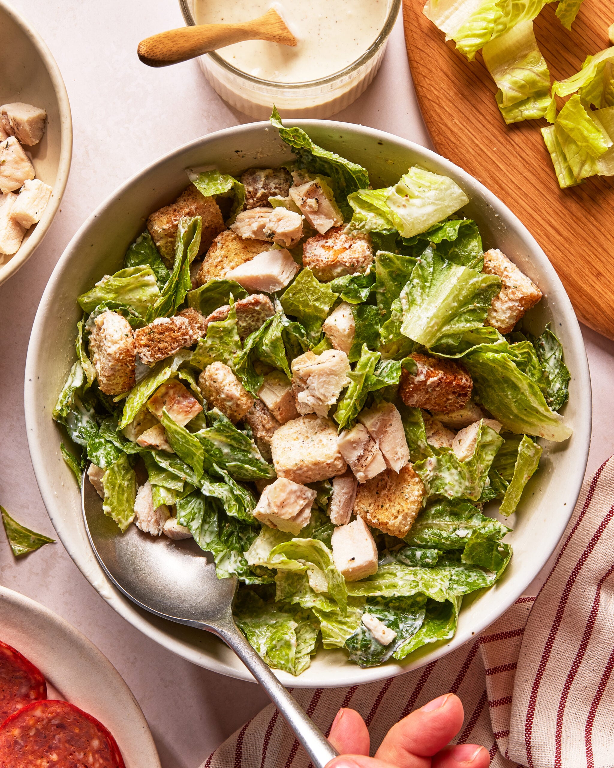 A bowl of Caesar salad with chopped romaine lettuce, grilled chicken pieces, croutons, and creamy dressing, with a spoon resting inside and a hand holding the bowl. Lettuce, dressing, and a striped napkin are nearby.