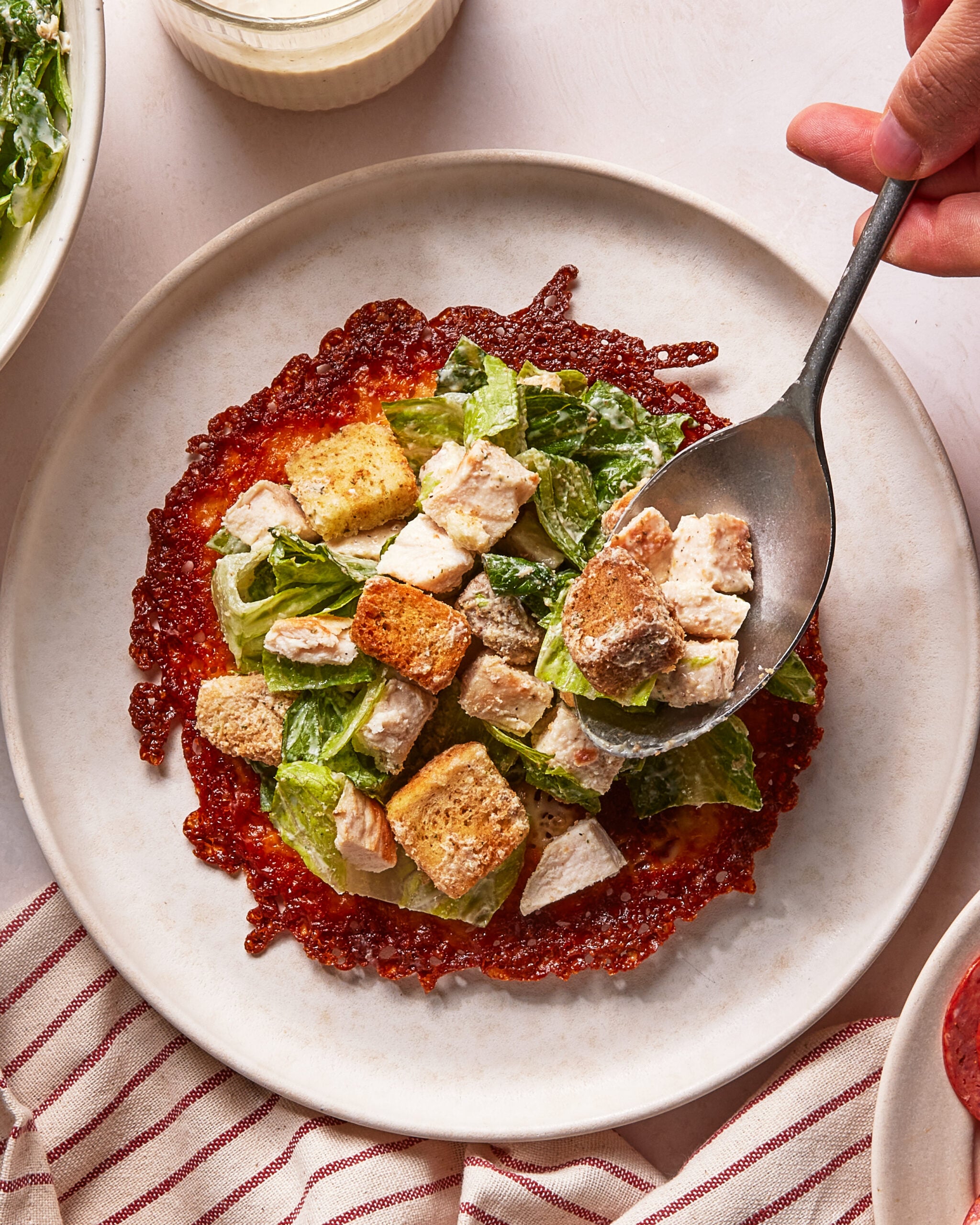 A plate with a crispy cheese base topped with romaine lettuce, croutons, and grilled chicken pieces, resembling a deconstructed Caesar salad. A hand holds a spoon, adding more chicken to the plate.