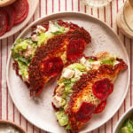 Two taco-shaped shells made from baked cheese and pepperoni, filled with chopped romaine lettuce and pieces of breaded chicken, are served on a white plate on a red-striped tablecloth. Bowls of ingredients surround the plate.