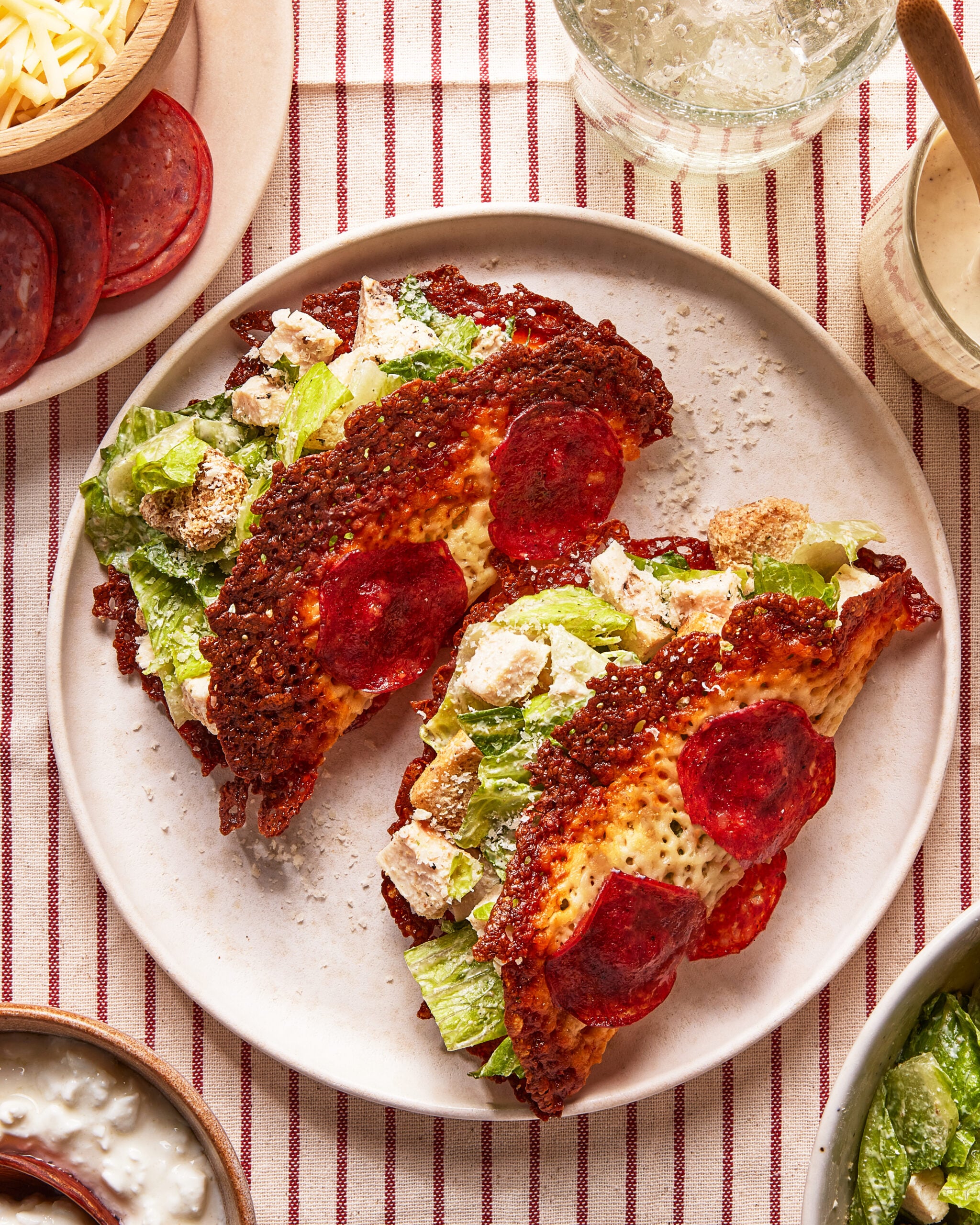 Two taco-shaped shells made from baked cheese and pepperoni, filled with chopped romaine lettuce and pieces of breaded chicken, are served on a white plate on a red-striped tablecloth. Bowls of ingredients surround the plate.