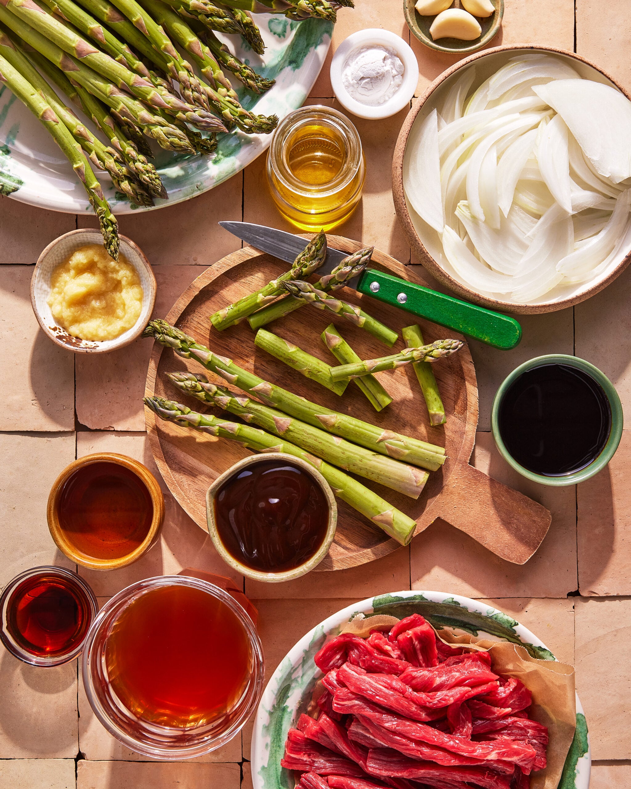 A top-down view of ingredients for a stir-fry: sliced beef, fresh asparagus, sliced onions, garlic, ginger, soy sauce, oil, and other sauces arranged on a tiled surface with a cutting board and knife.