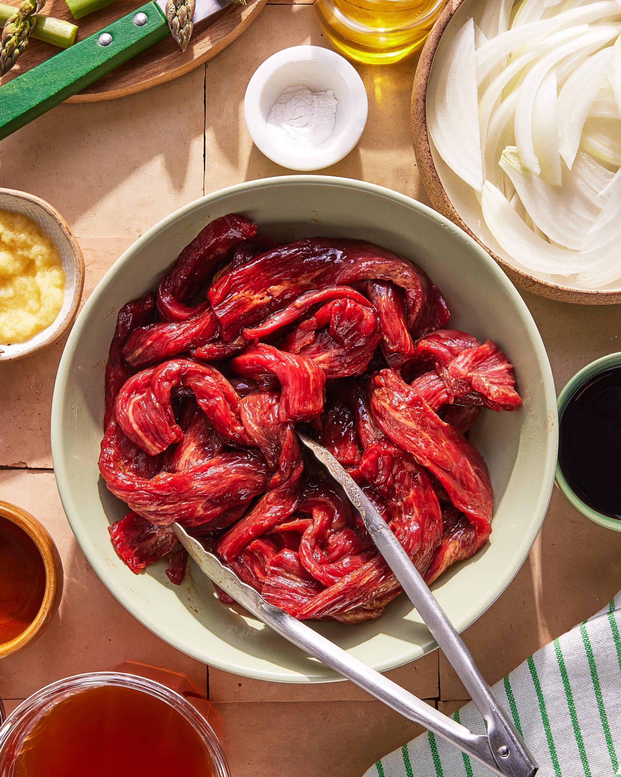 A bowl of raw marinated beef strips with metal tongs, surrounded by sliced onions, sauces, oil, a knife with asparagus, and a striped towel on a tiled surface.