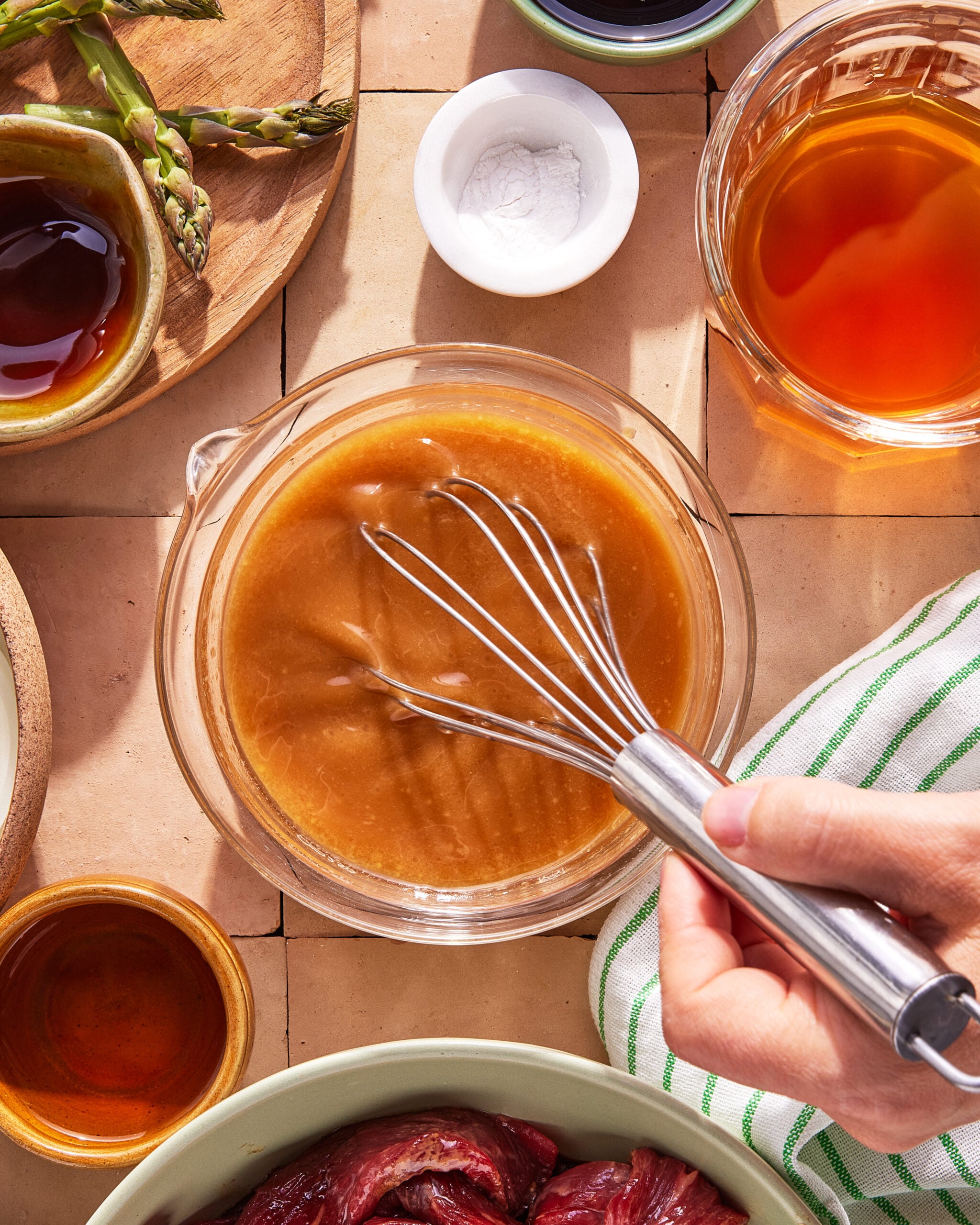A hand holds a whisk, mixing a brown sauce in a glass bowl on a tiled surface. Surrounding the bowl are ingredients like asparagus, small bowls of liquid, and a green-striped towel.