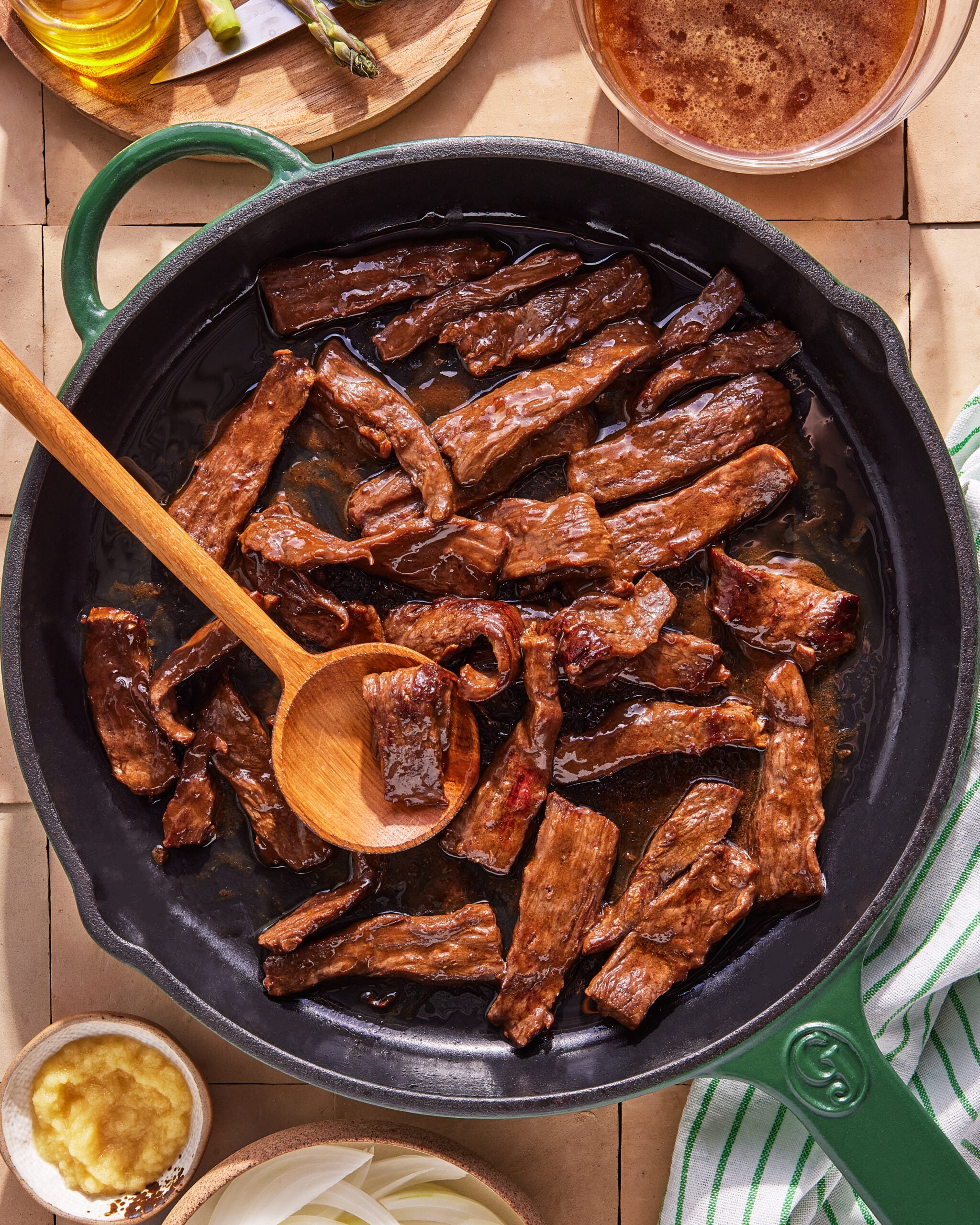 A cast iron skillet filled with cooked strips of beef being stirred with a wooden spoon, surrounded by small bowls of sauce, garlic paste, and sliced onions on a tiled surface.