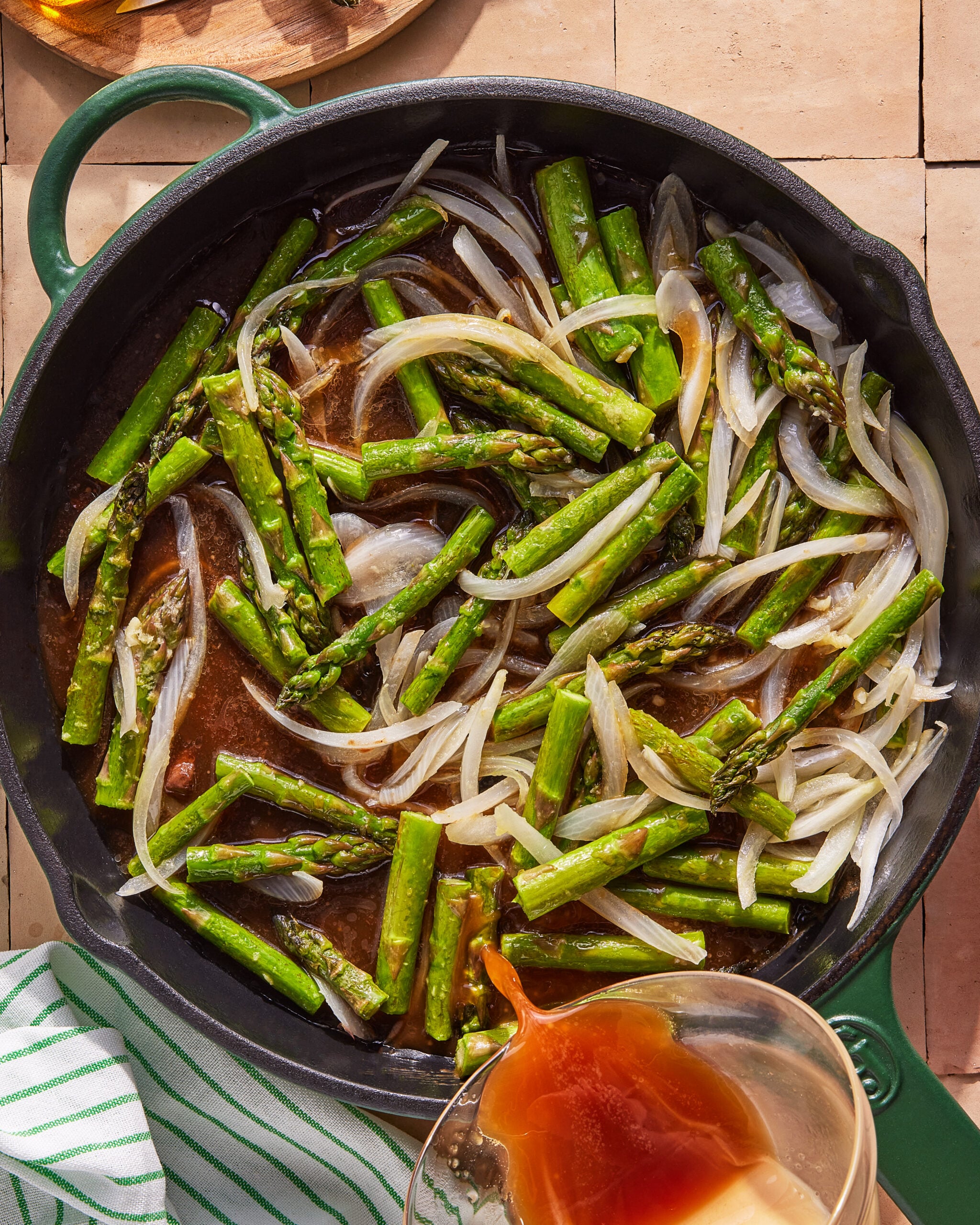 A cast iron skillet filled with sautéed asparagus and sliced onions, with a hand pouring sauce or broth from a glass measuring cup into the pan. A green and white striped towel is nearby.
