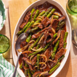 Oval platter with beef stir-fry featuring strips of beef, asparagus, and onions in a brown sauce, served with a spoon. Green drinks and striped napkin are nearby on a wooden table.