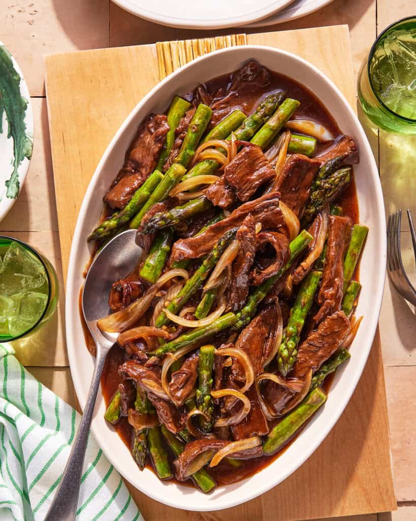 Oval platter with beef stir-fry featuring strips of beef, asparagus, and onions in a brown sauce, served with a spoon. Green drinks and striped napkin are nearby on a wooden table.