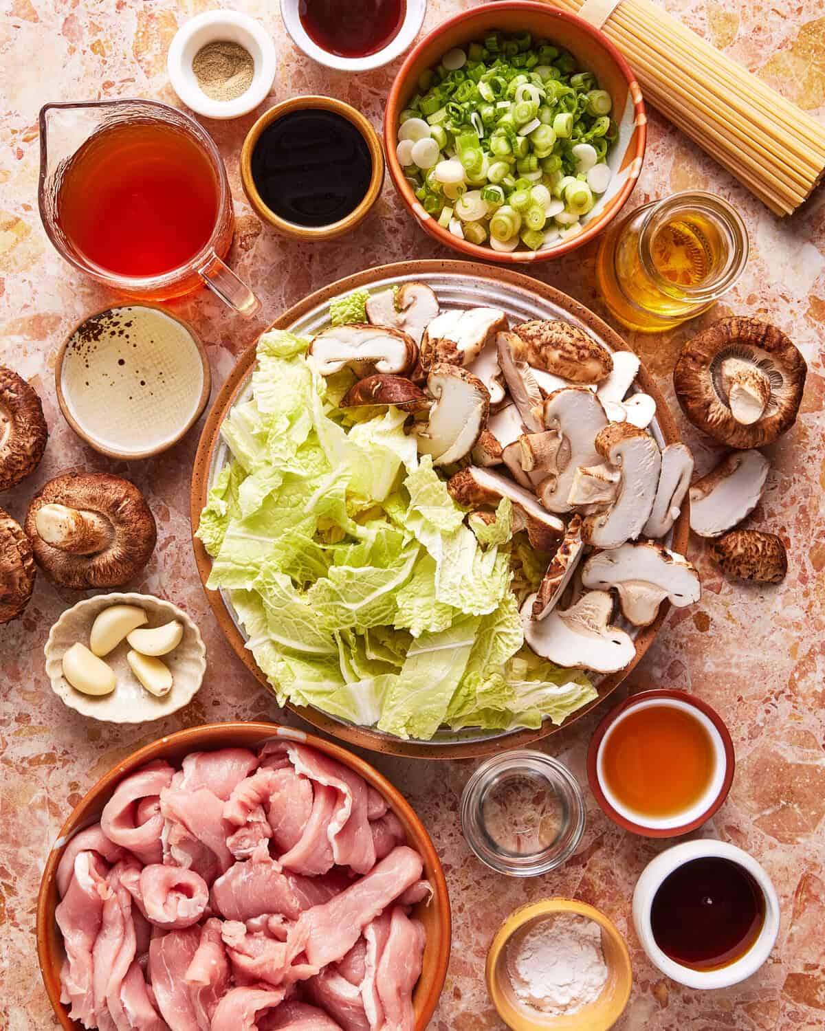 A top view of various ingredients for an Asian hot pot, including sliced pork, napa cabbage, shiitake mushrooms, green onions, garlic, noodles, and small bowls of sauces and seasonings on a marble surface.