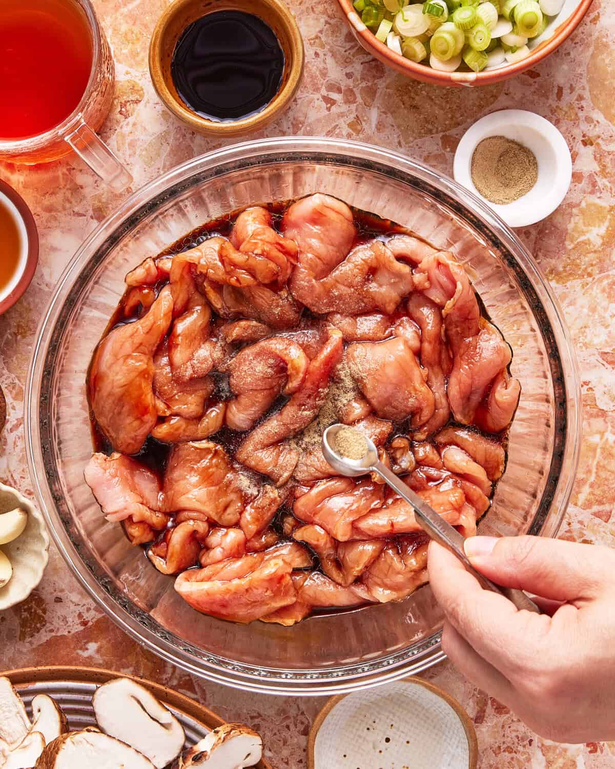 A hand sprinkles pepper onto raw marinated meat in a glass bowl surrounded by ingredients such as soy sauce, green onions, broth, mushrooms, and other seasonings on a marble countertop.